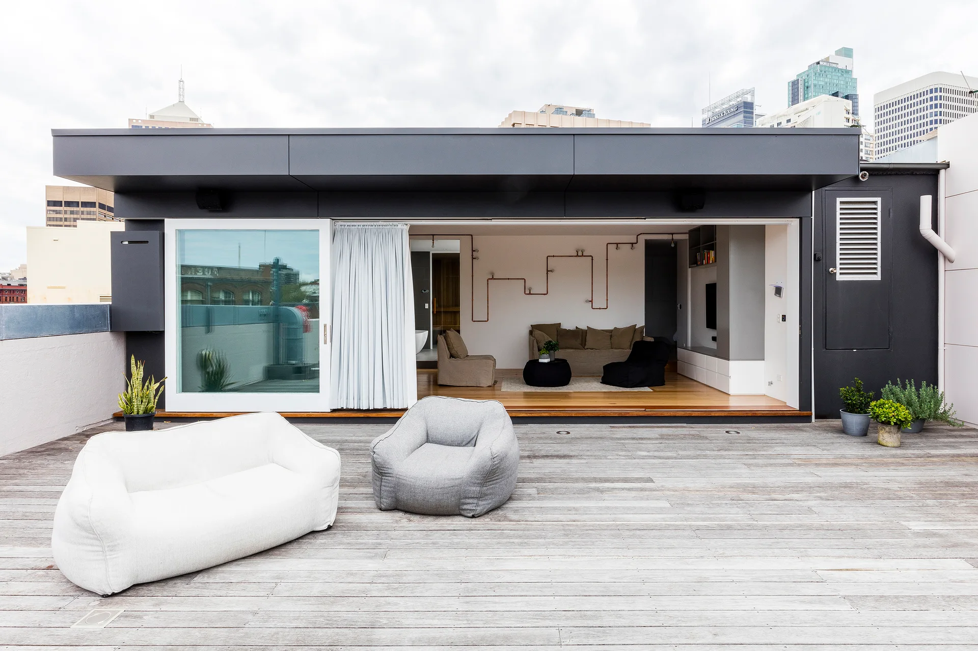 A spacious rooftop wood deck with casual grey and white lounge chairs transitions through a wide glass door into an indoor living room.