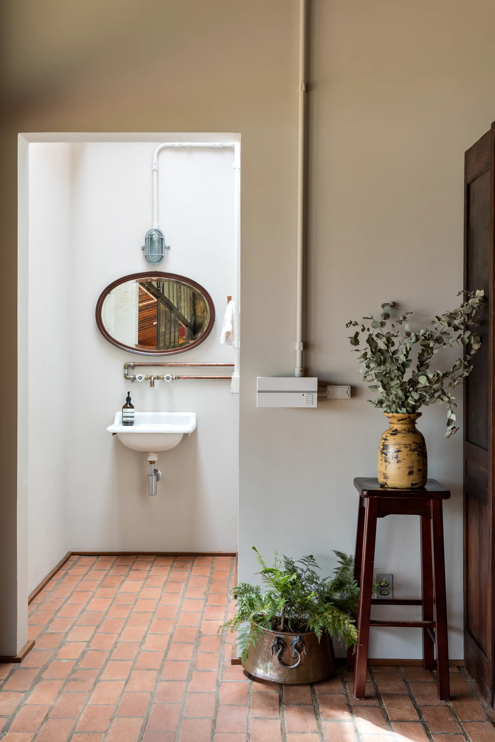 A rustic bathroom view showing terracotta brick flooring, a small wall-mounted sink with exposed copper pipes, and an oval mirror.