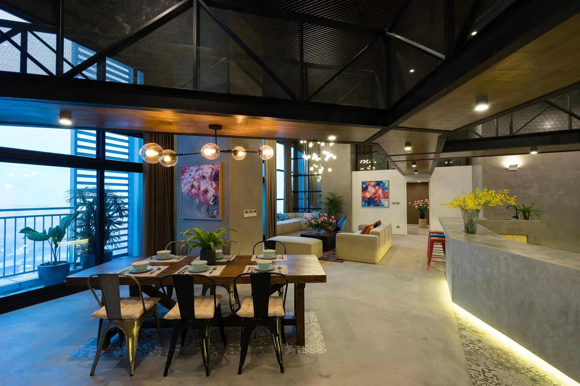 An industrial open-concept dining room features a rustic wooden table, metal chairs, a concrete floor with a patterned tile inlay, and exposed ceiling beams.