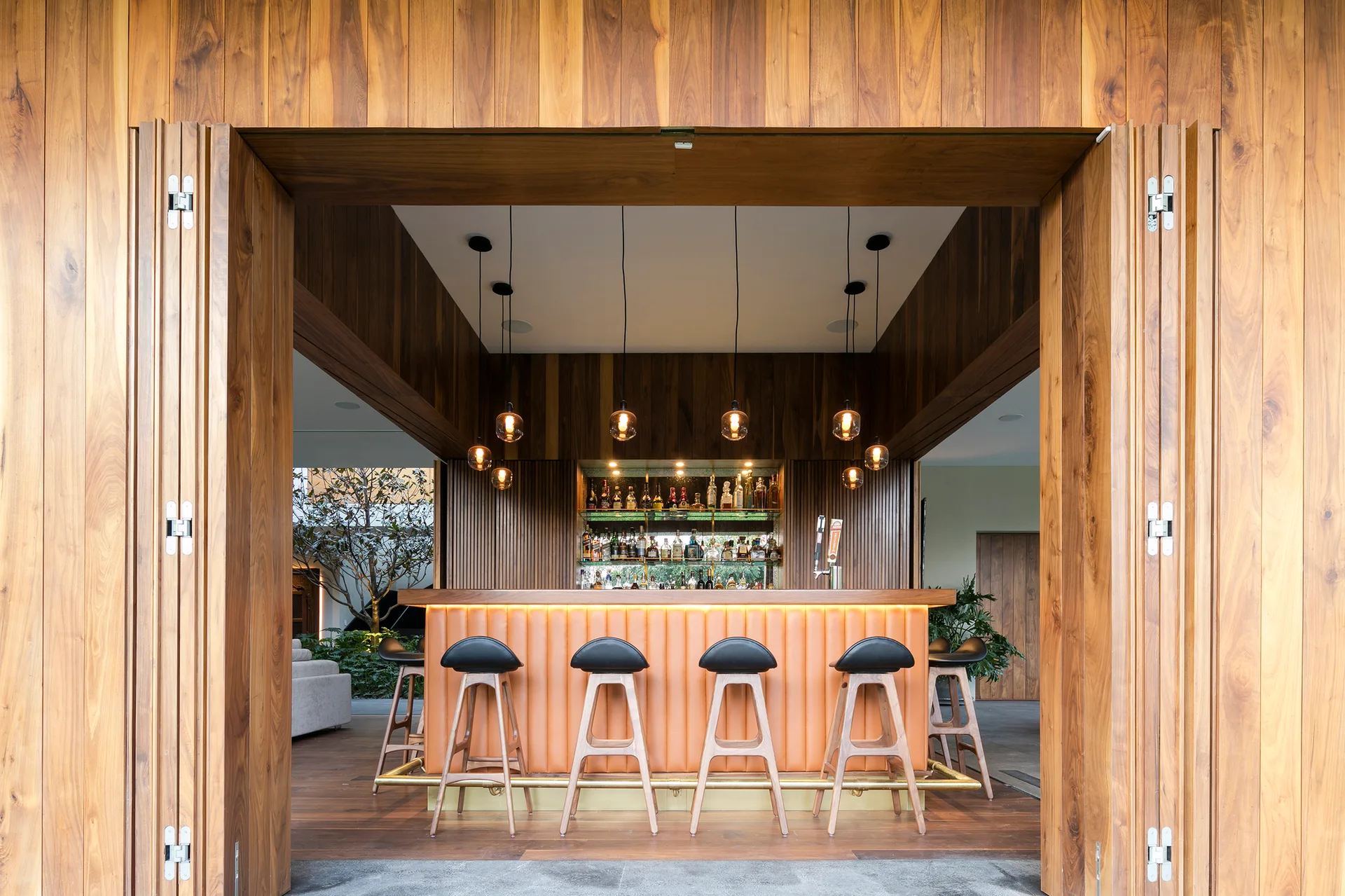 An indoor bar framed by tall wooden bifold doors features a fluted orange counter, wood bar stools, and delicate hanging pendant lights.
