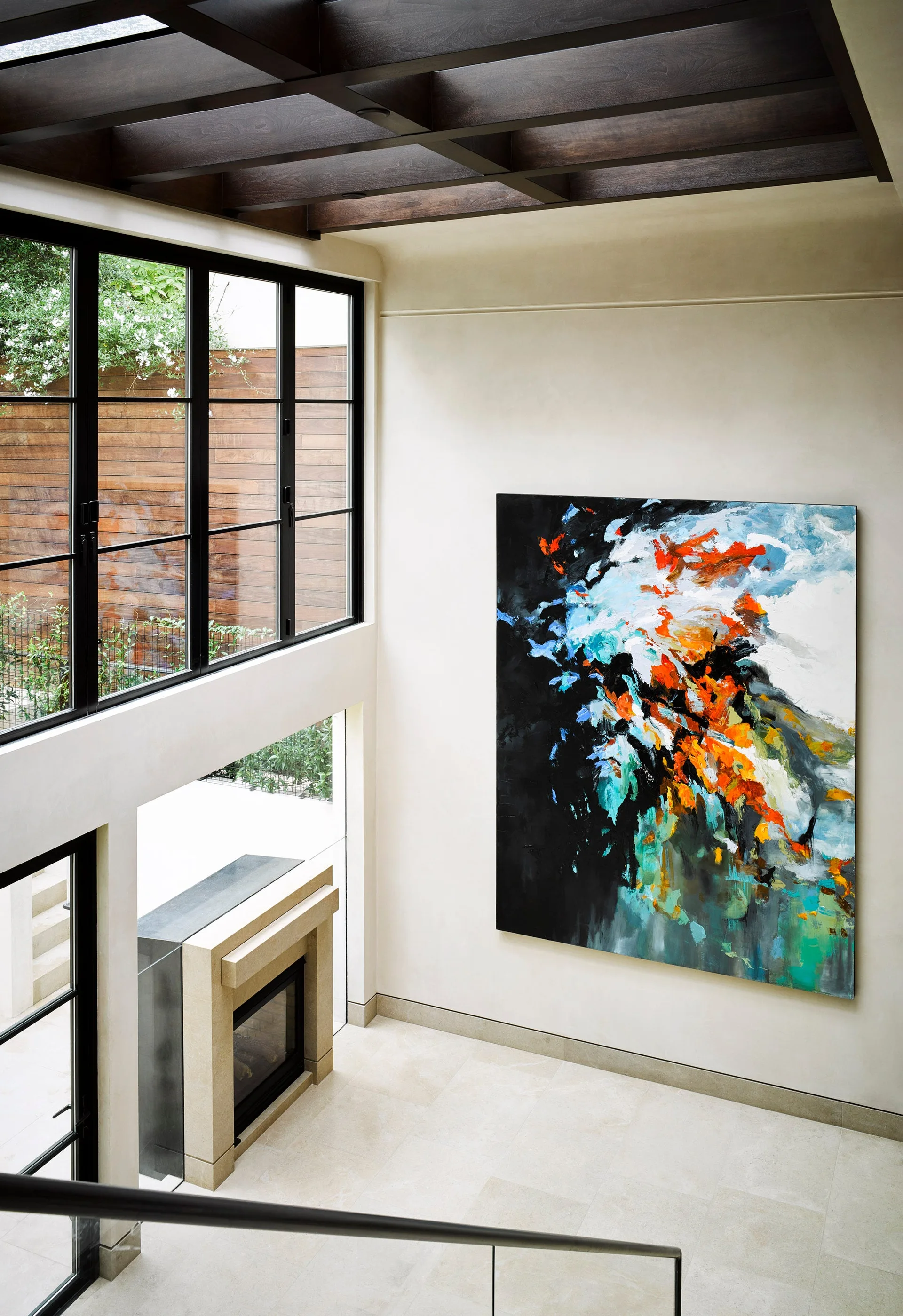 A high-ceilinged room with a massive black and orange abstract painting on a cream wall next to large gridded windows.