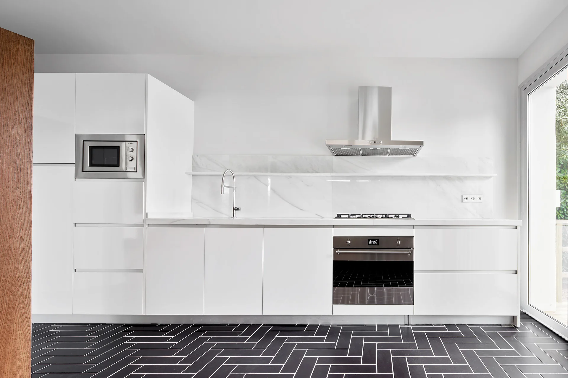 A modern white kitchen with high-gloss cabinetry, a slab marble backsplash, and a dark herringbone tile floor.
