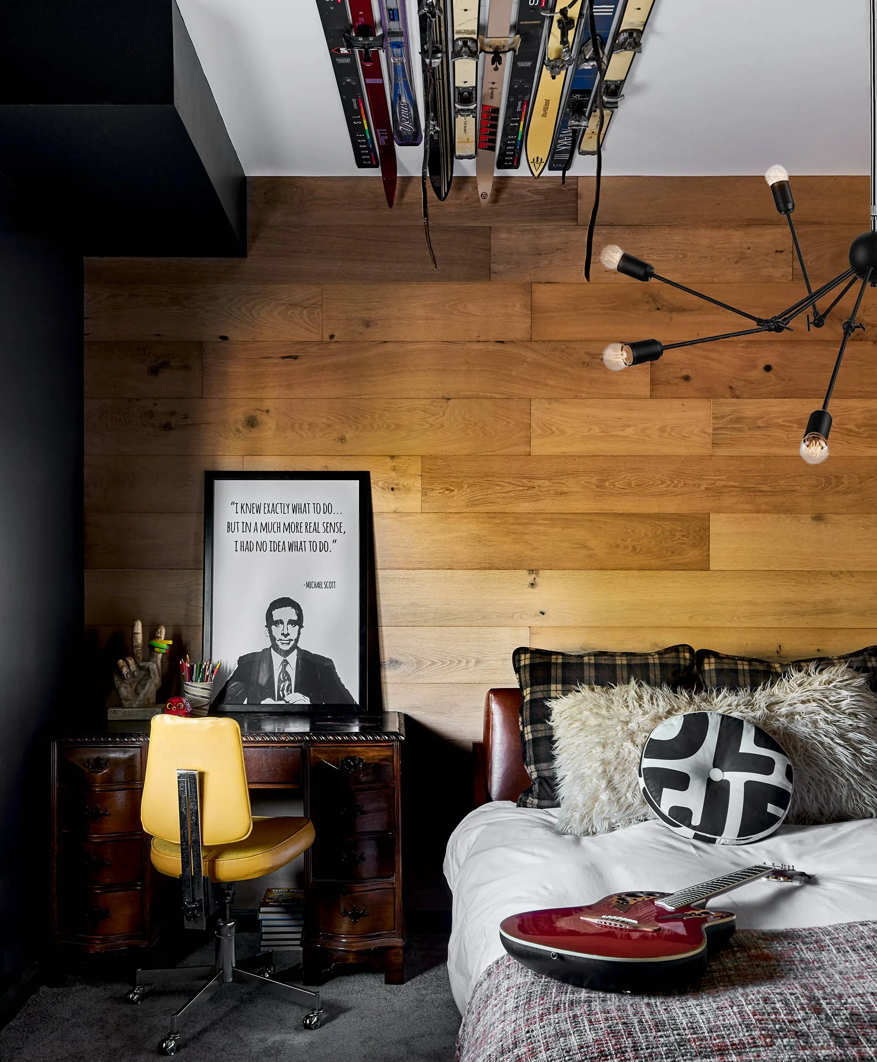 A bedroom features horizontal wood wall paneling, an antique desk with a yellow swivel chair, and vintage skis mounted near the white ceiling.