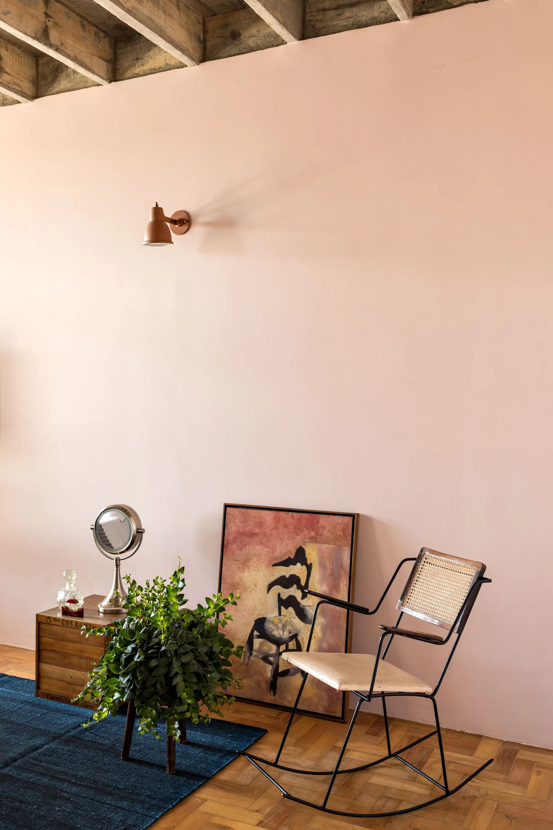 A cane-backed rocking chair sits on parquet flooring beside a framed painting resting on the floor against a pastel pink wall.