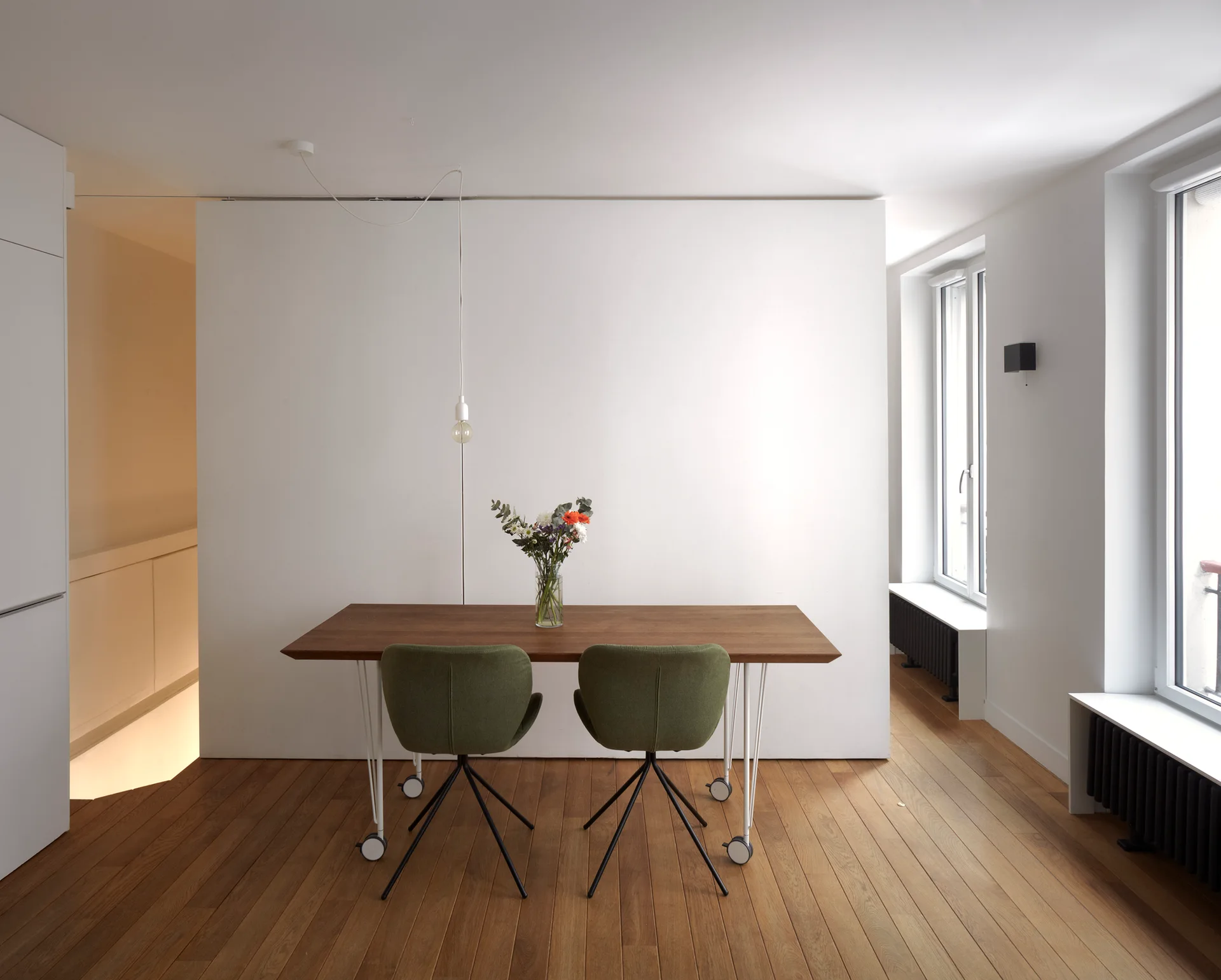 A wooden dining table on caster wheels is paired with two olive green chairs in front of a solid white partition wall.