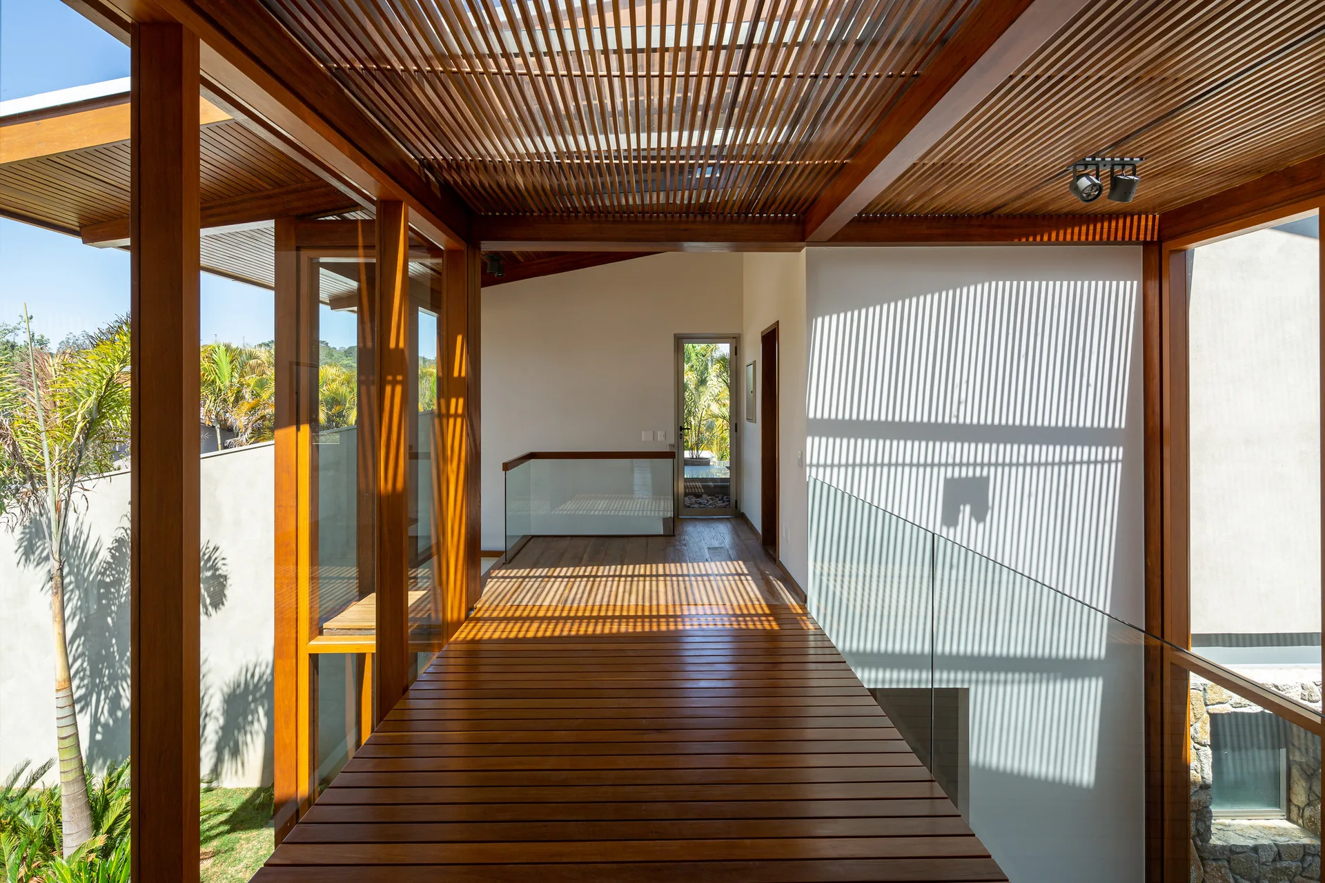 A modern indoor wooden walkway features a slatted ceiling, glass railings, white walls with linear shadows, and outdoor views.