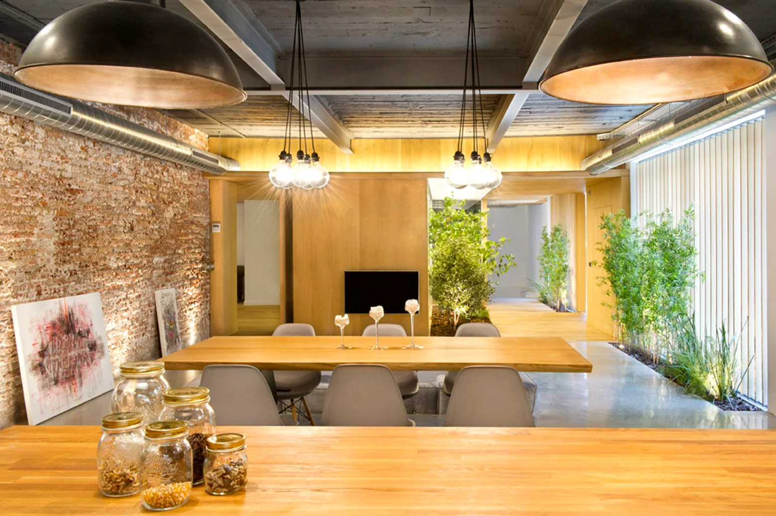 A modern industrial dining room featuring a long wooden dining table, grey chairs, an exposed brick wall, and large dome pendant lights.