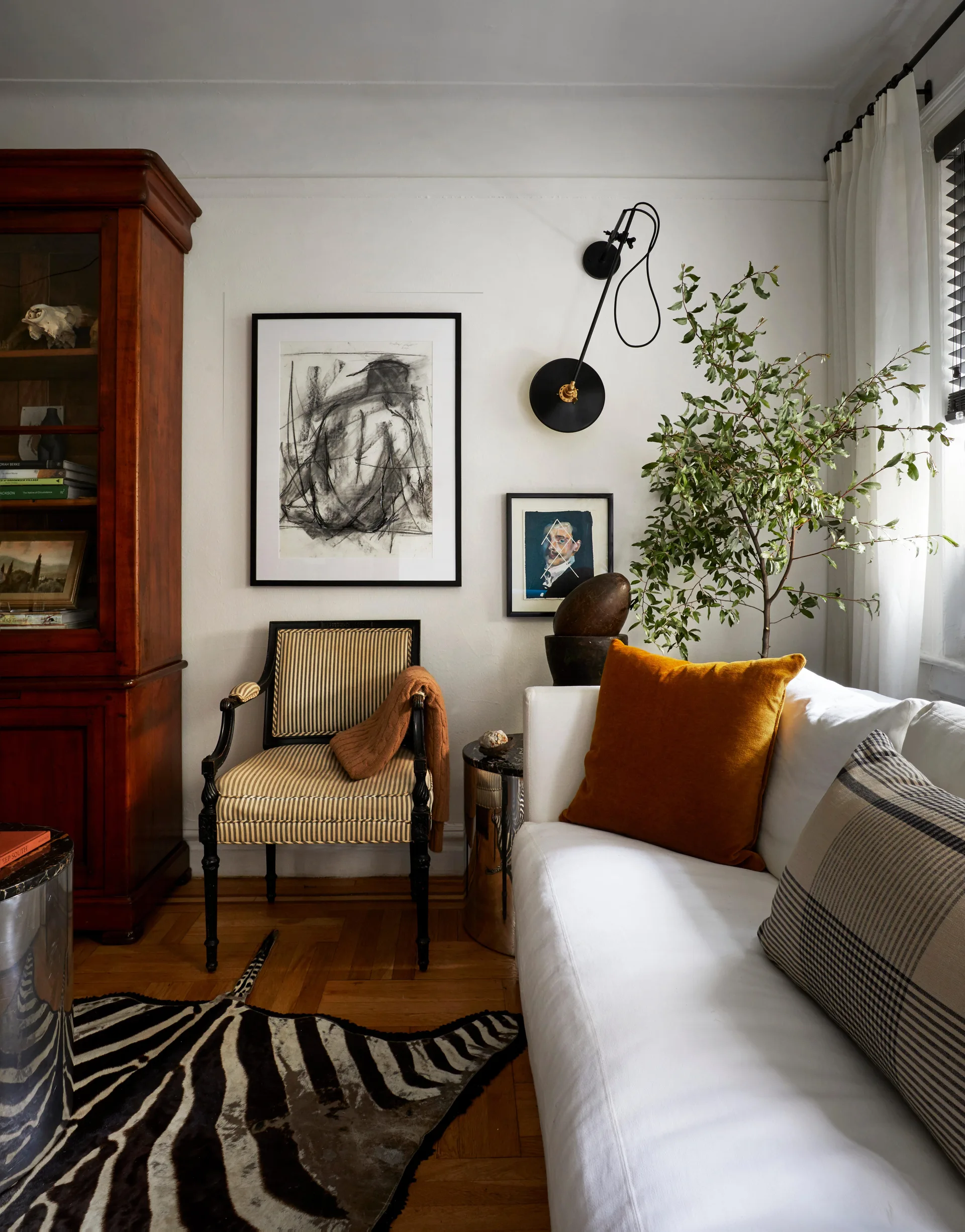 A decorated living room corner features a white sofa, a striped armchair, a tall wooden cabinet, framed artwork, and a zebra-print hide rug.