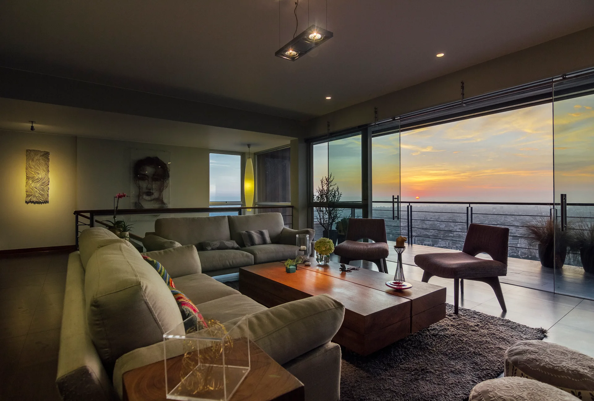 A modern living room features gray sofas, a low block wood coffee table, and floor-to-ceiling glass doors opening to a city sunset view.