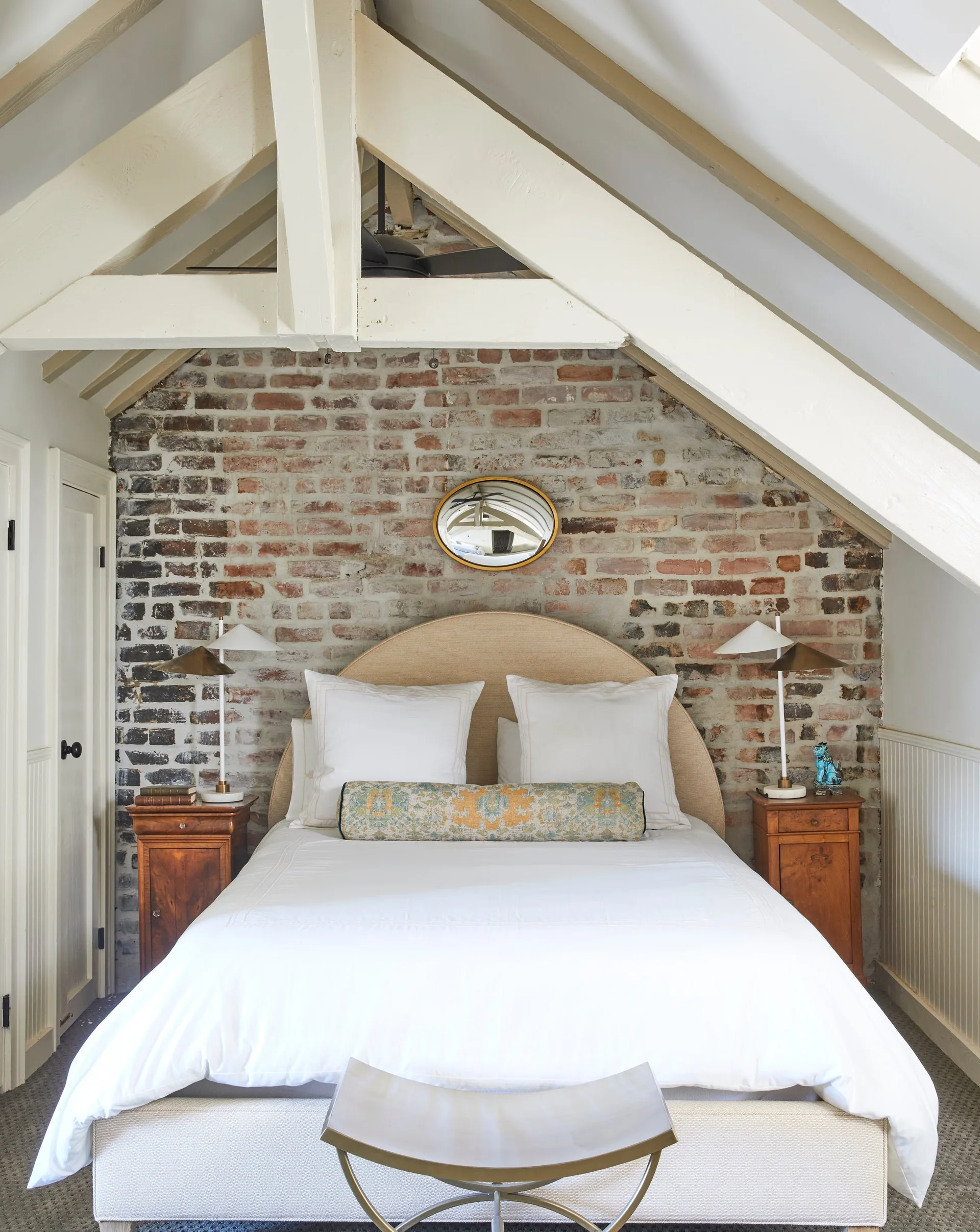 An attic bedroom features an upholstered bed with white linens against a rustic exposed brick wall beneath sloped white ceilings and beams.