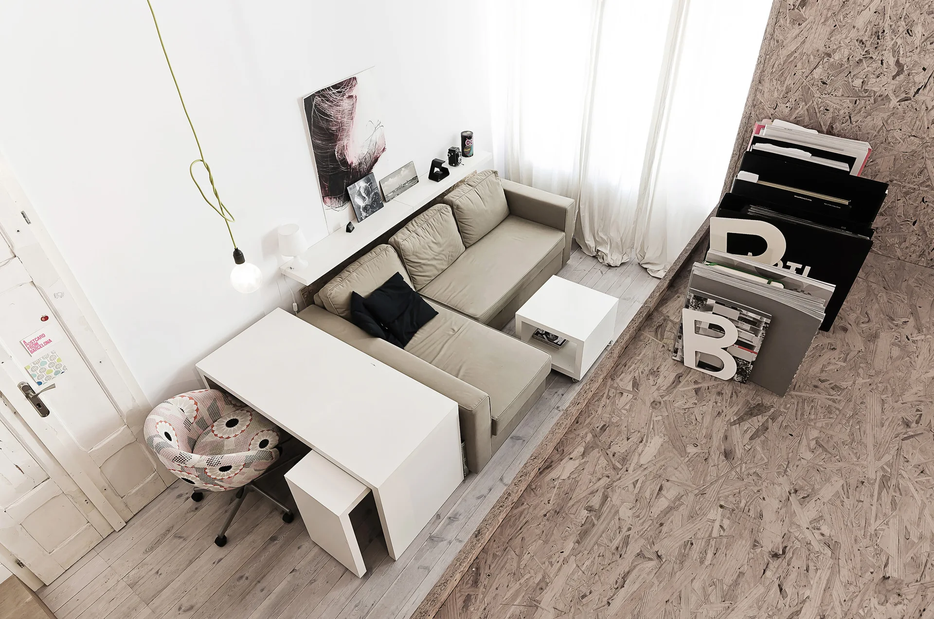 An overhead view of a modern studio apartment showing a tan sectional sofa, a white desk with a patterned chair, and oriented strand board flooring.