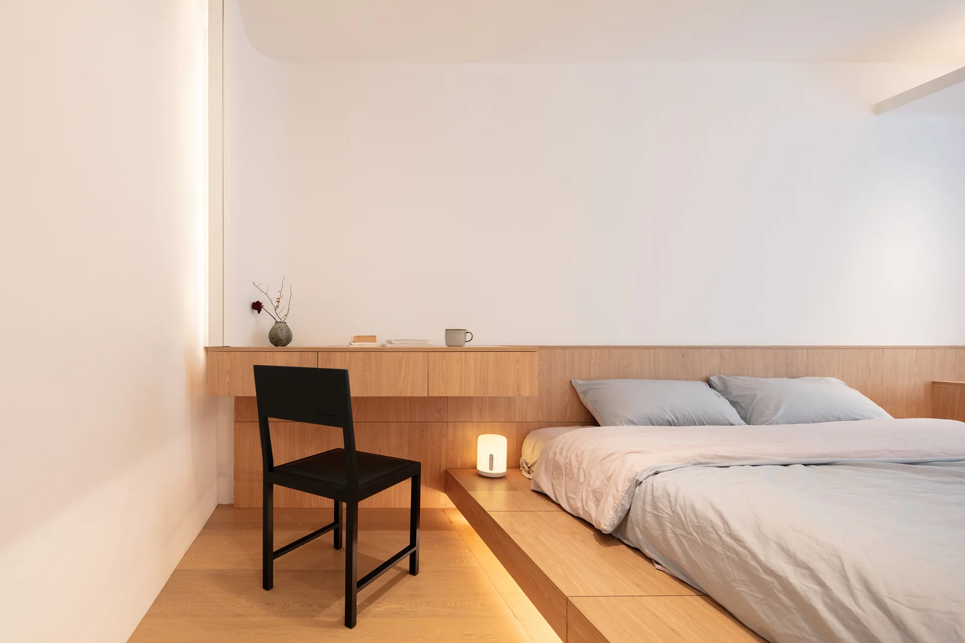 A minimalist bedroom featuring a light wood platform bed, an integrated floating desk, warm under-bed lighting, and a single black chair.