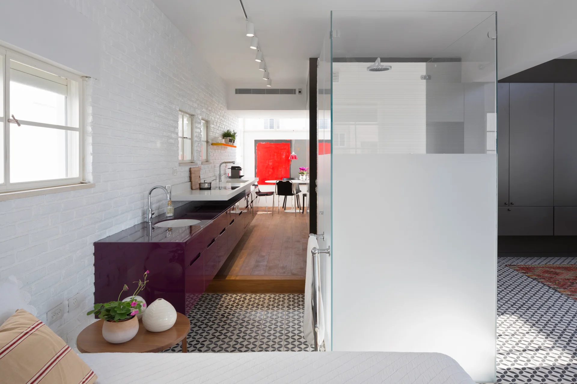 An open modern bathroom features a long glossy purple vanity against a white painted brick wall next to a glass-enclosed shower and geometric floor tiles.