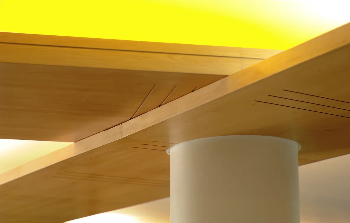 A close-up of intersecting light wood ceiling panels with recessed grooves meeting above a round white column under a bright yellow light glow.