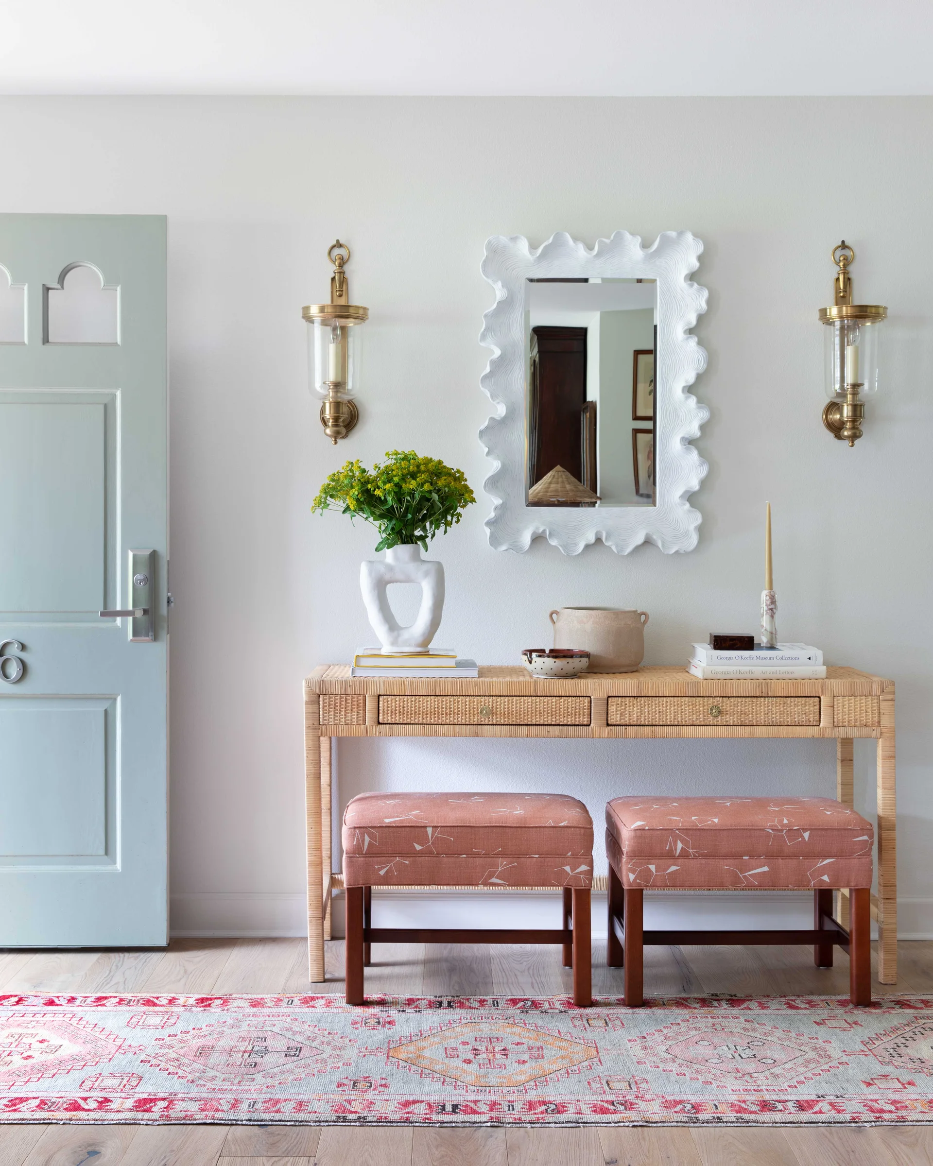 An entryway features a rattan console table with two terracotta ottomans underneath, flanked by a light blue door and topped with a wavy white mirror.
