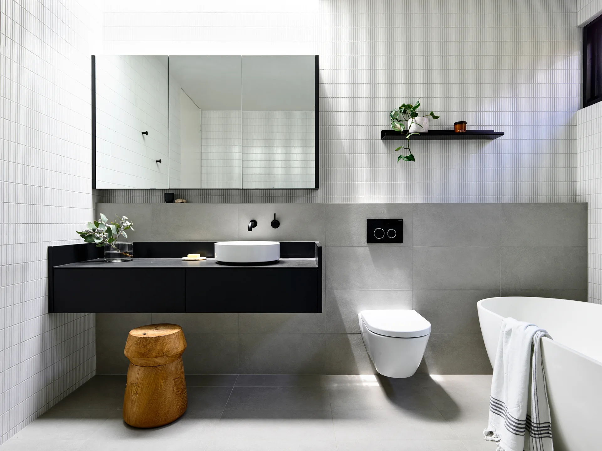 A modern bathroom featuring a black floating vanity with a white vessel sink, a wall-mounted toilet, and slim vertical white wall tiles.
