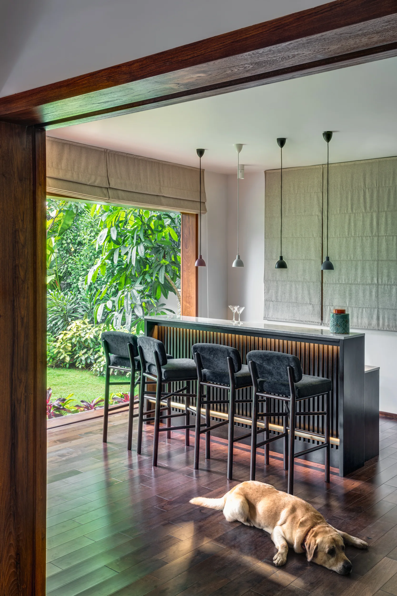 A modern home bar with four dark stools features a slatted wood front and under-counter lighting, set against a large window with a garden view.