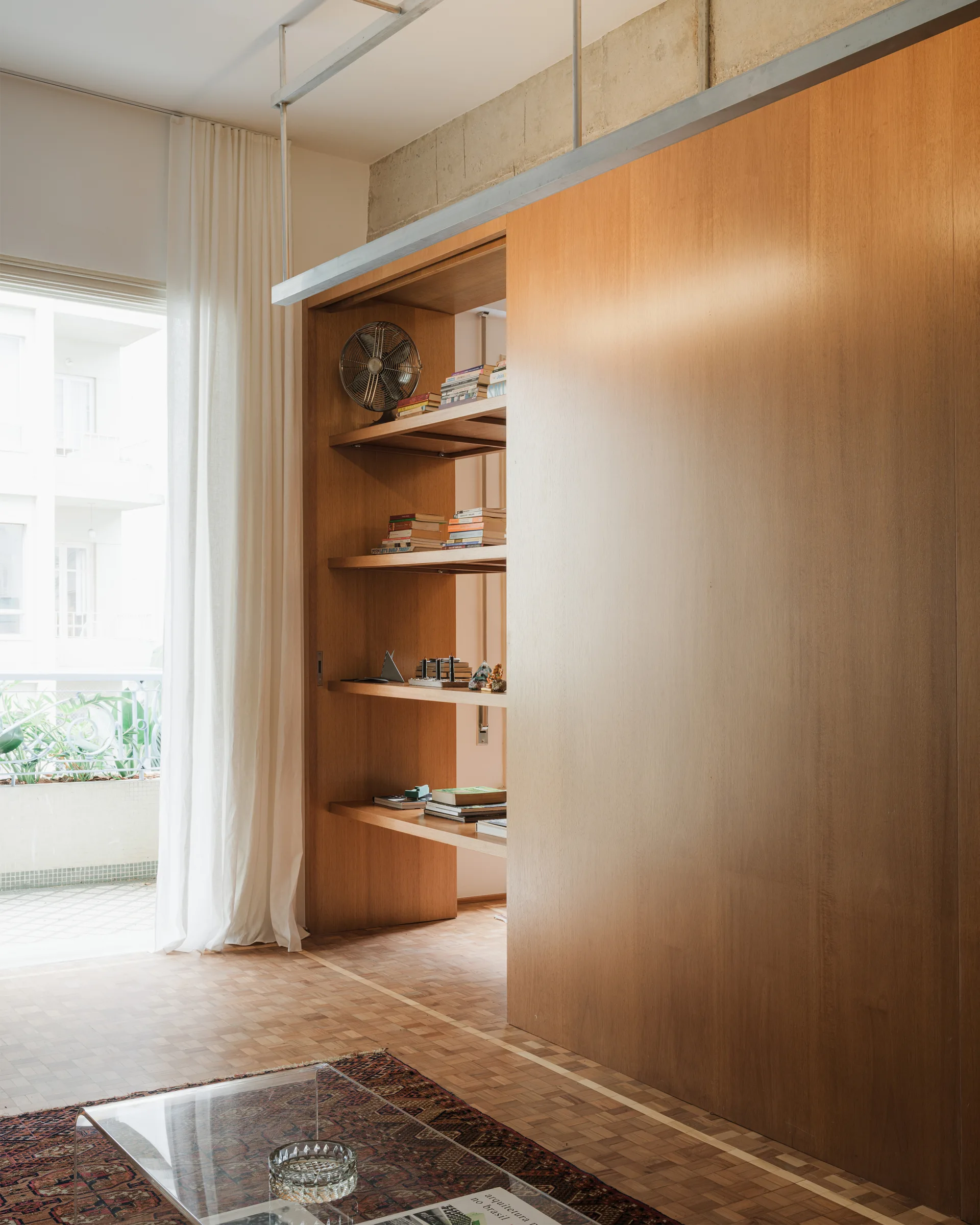 A large sliding wood partition wall with built-in shelves stands on parquet flooring next to a balcony window with white sheer curtains.