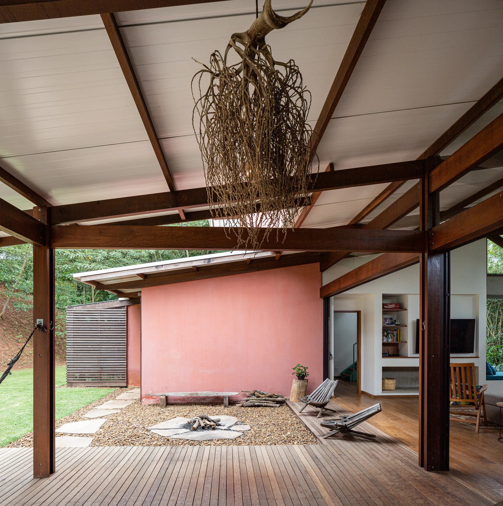 A covered wooden patio features a gravel-lined fire pit, slatted lounge chairs, a terracotta accent wall, and a sculptural organic pendant light.