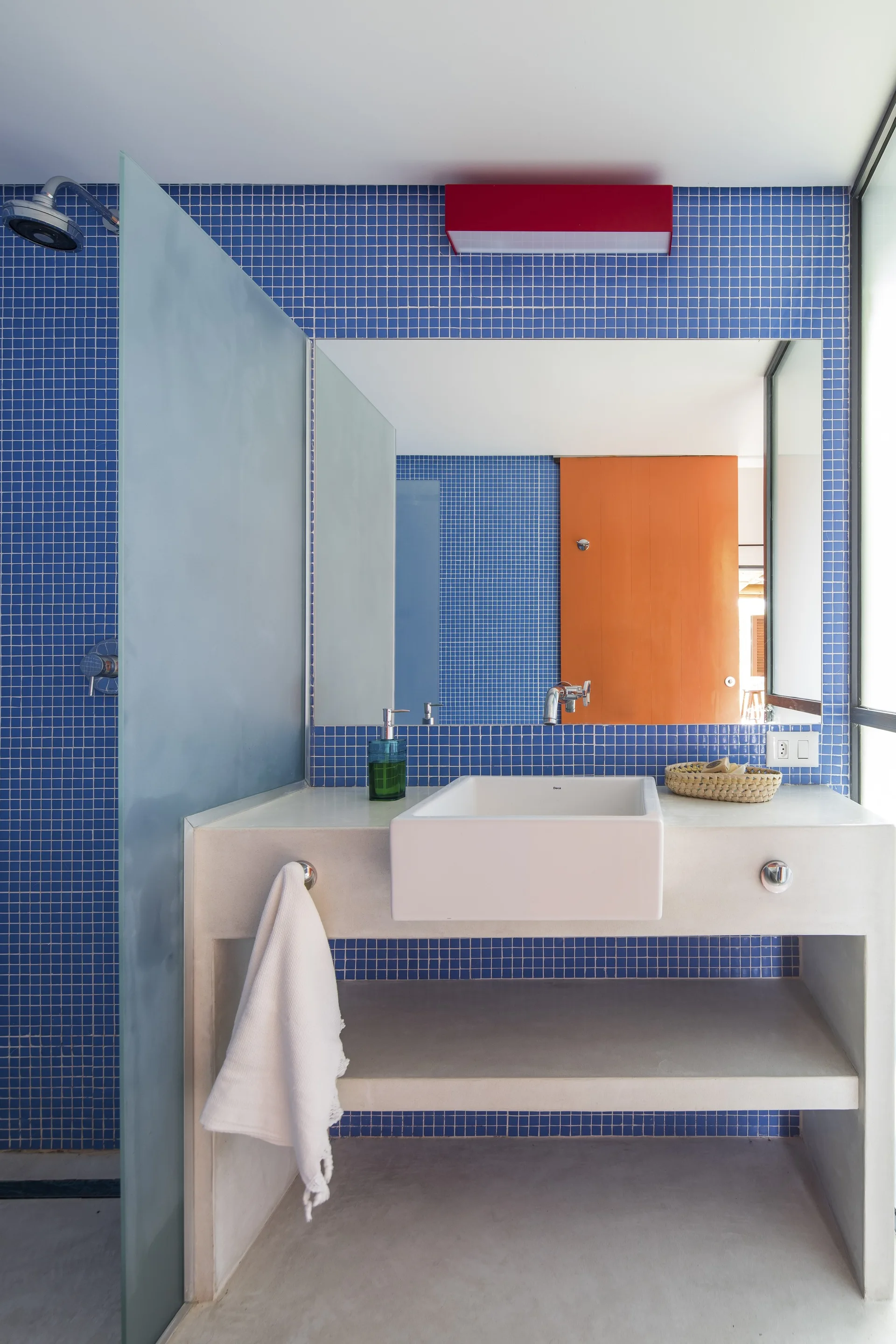 A modern bathroom features blue mosaic wall tiles, a light gray concrete vanity with a white basin sink, and a mirror reflecting an orange door.