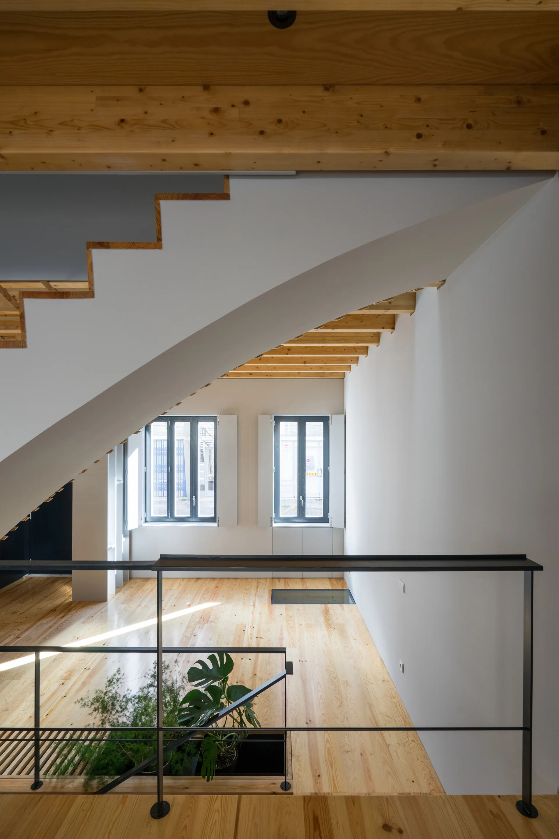 A modern interior features a white diagonal staircase, light wood flooring, exposed timber ceiling beams, and simple black metal railings.