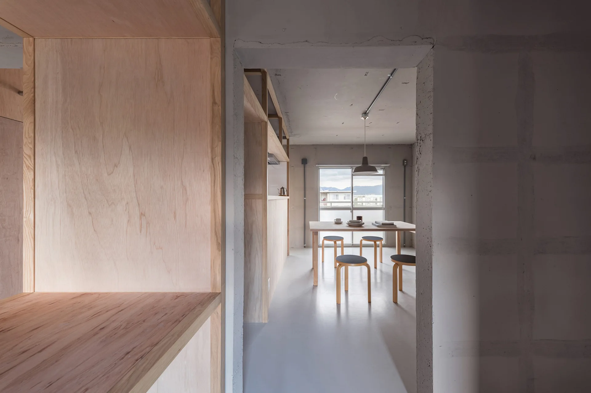 A view through a concrete doorway reveals a minimalist dining room with a light wood table, four round stools, and custom plywood shelving.