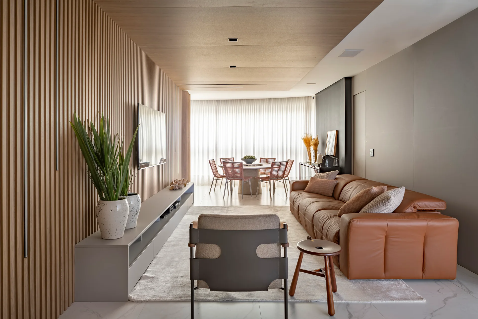 A modern living room features a vertical wood slat wall, a brown leather sofa, and a dining area with sheer curtains in the background.