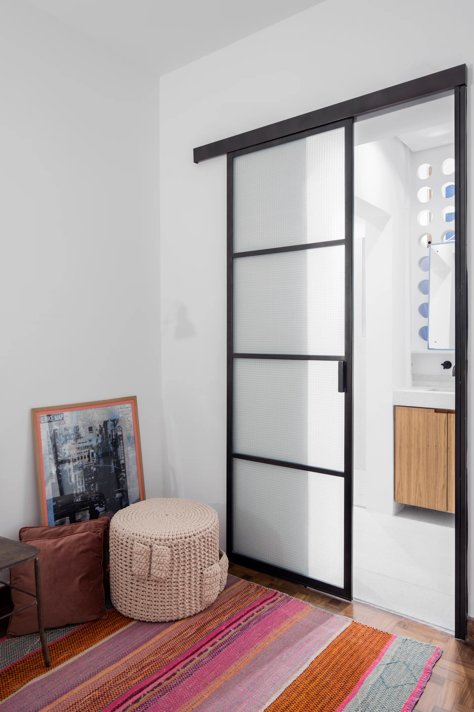 A black-framed sliding door with frosted grid glass panels separates a colorful bedroom corner from a bright bathroom vanity area.