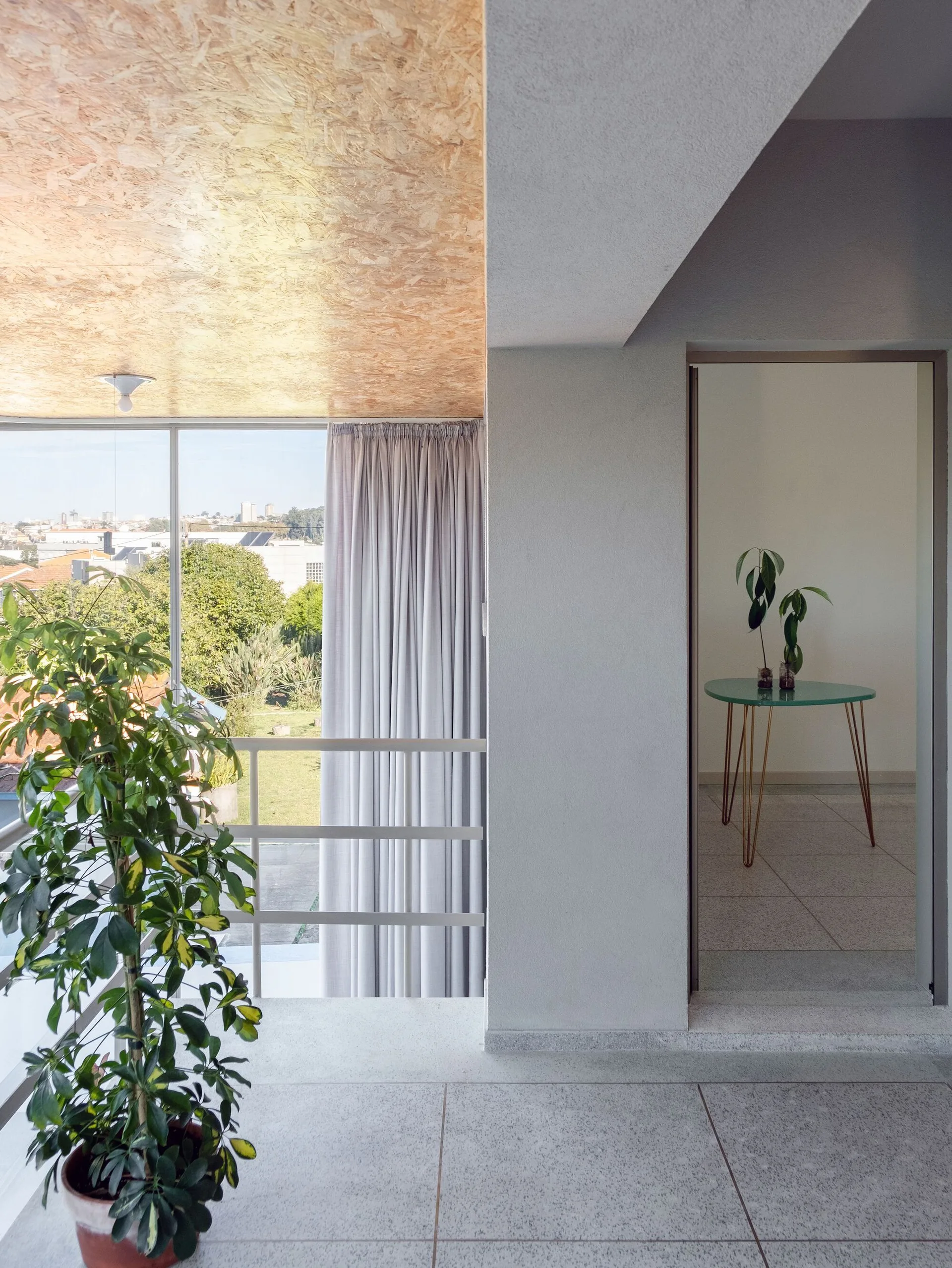 A modernist room features a textured OSB ceiling, light grey walls, a balcony window with drapes, and a doorway looking into a side table.