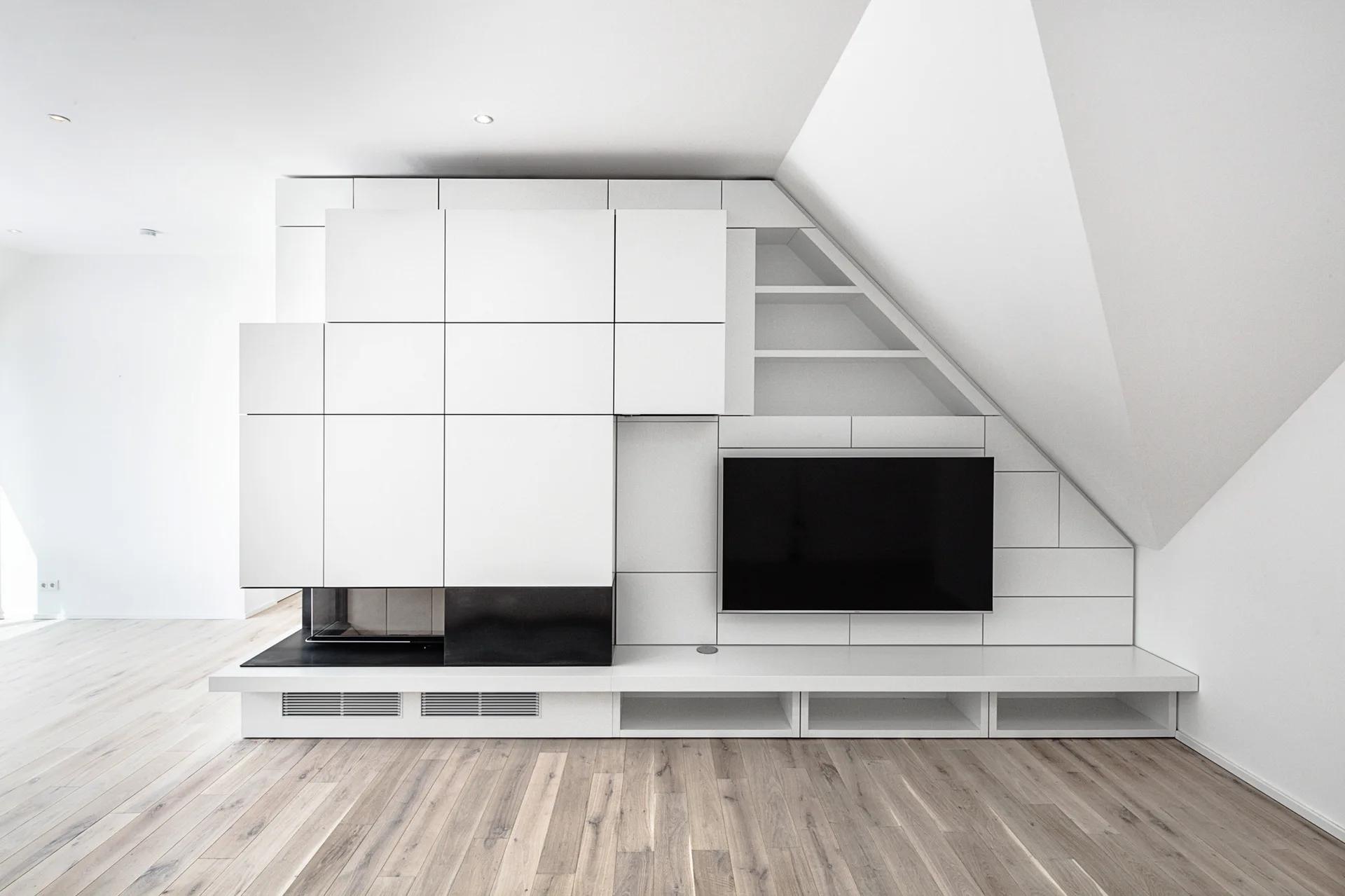 A custom white minimalist media wall featuring an integrated television and fireplace sits beneath a sloped ceiling on light wood floors.