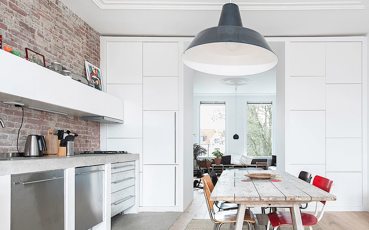 An industrial kitchen features an exposed brick wall, white integrated cabinets, and a rustic wood dining table with mismatched vintage chairs.