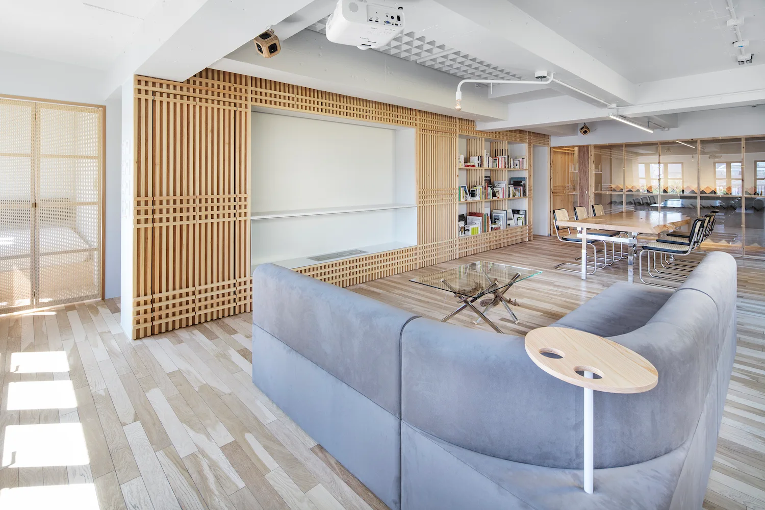 A bright modern living room features a grey sectional sofa, light wood floors, and a large wooden slatted wall unit with built-in shelves.