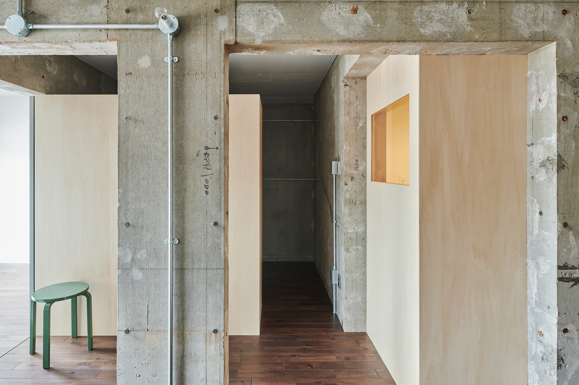 A minimalist interior featuring raw concrete structural beams, light plywood partition walls, exposed metal conduits, and a green stool.