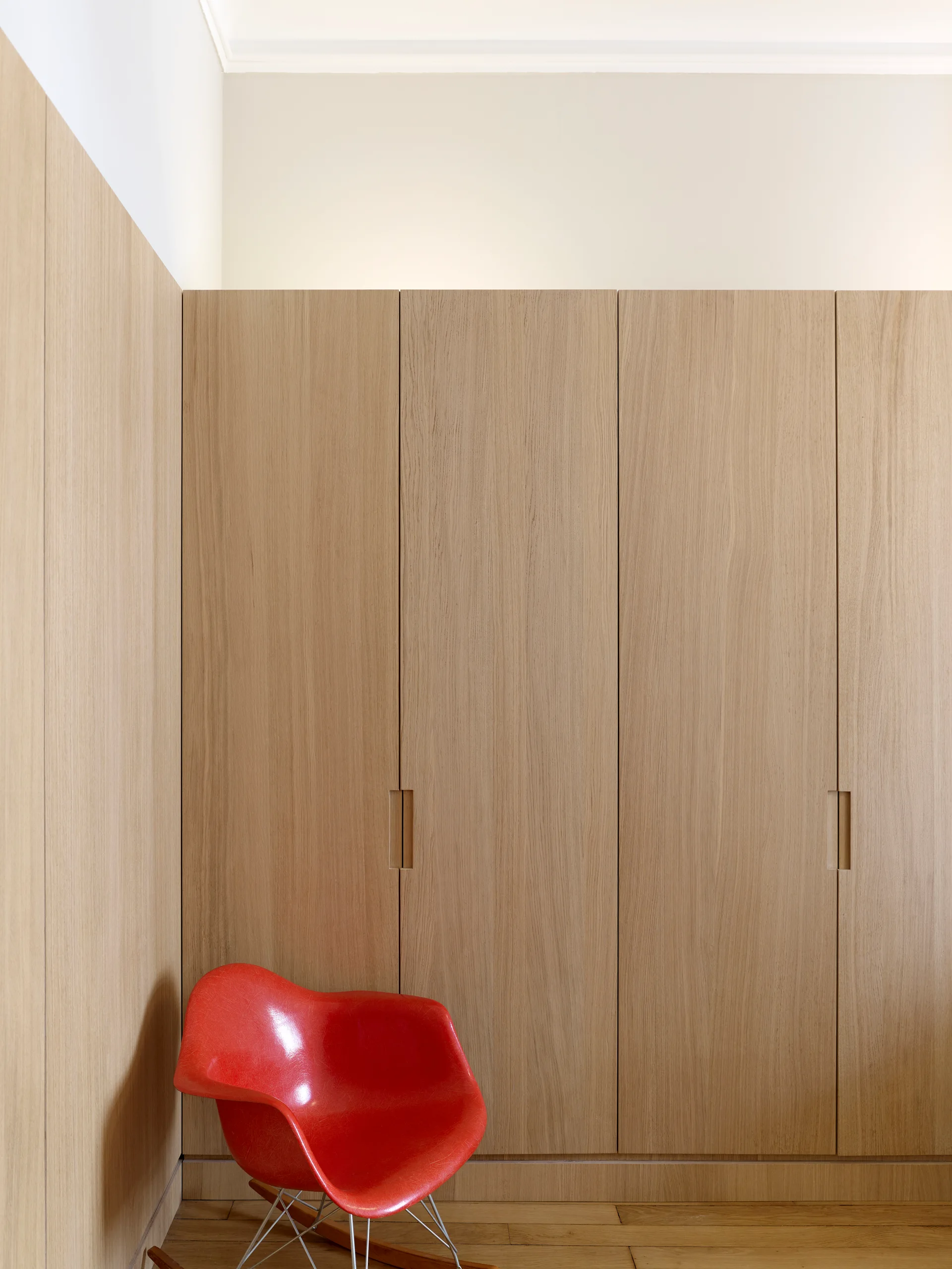 A red molded plastic chair sits on a wood floor in front of a floor-to-ceiling light oak wood paneled wardrobe closet.
