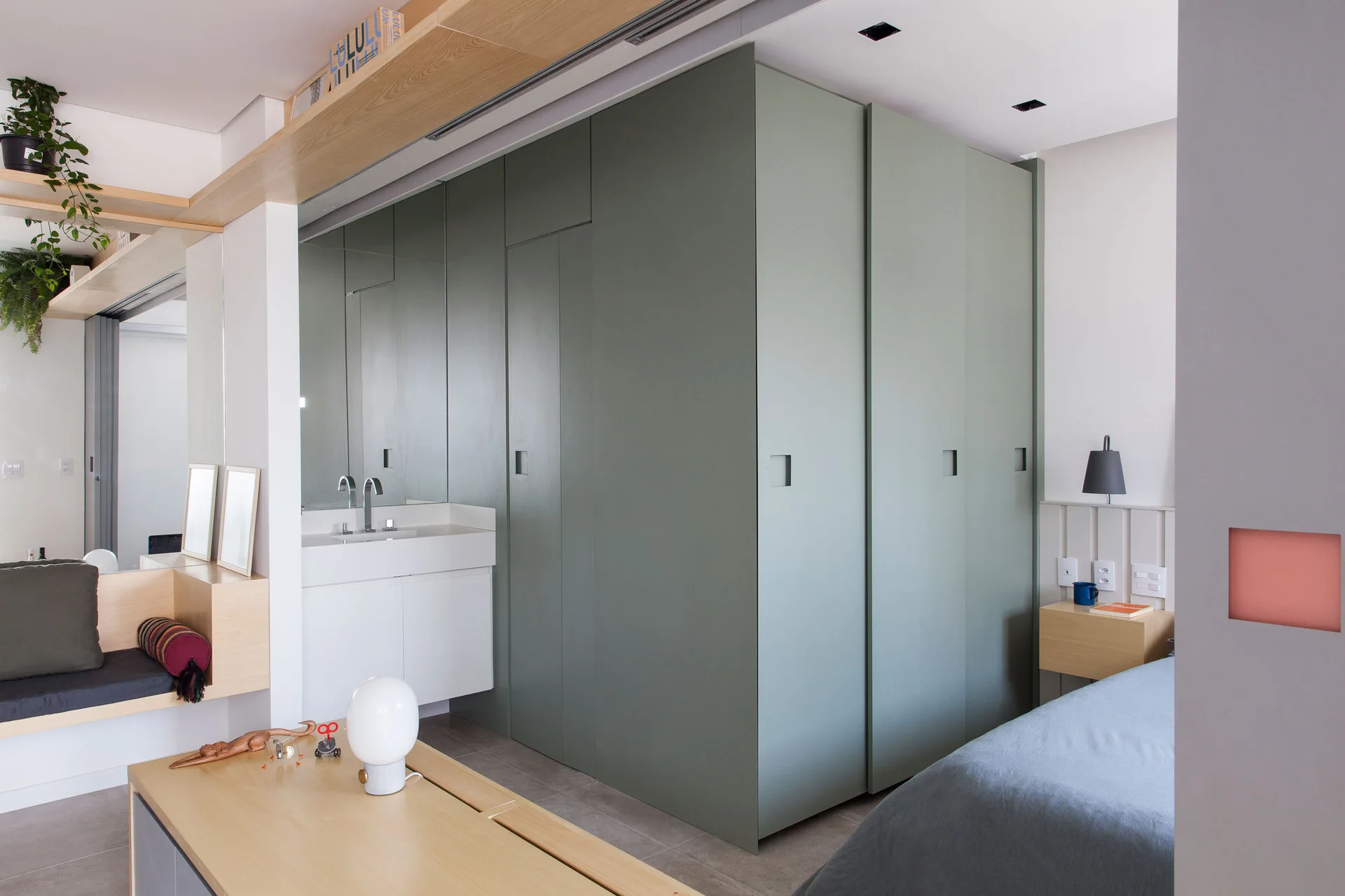 A modern bedroom featuring a large sage green built-in wardrobe, an integrated white vanity sink with a mirror, and a light wood floating nightstand.