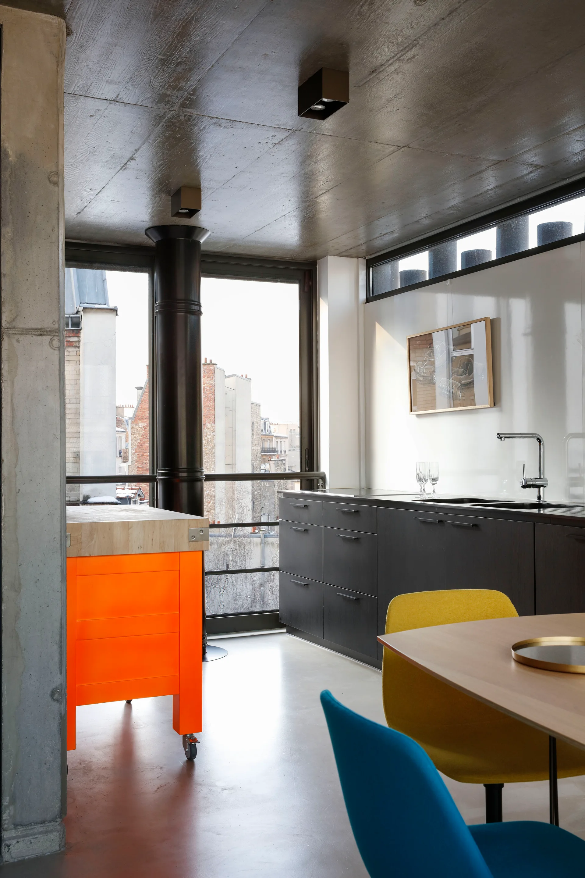 A modern kitchen features dark gray cabinets, an exposed concrete ceiling, a bright orange rolling island, and colorful dining chairs.
