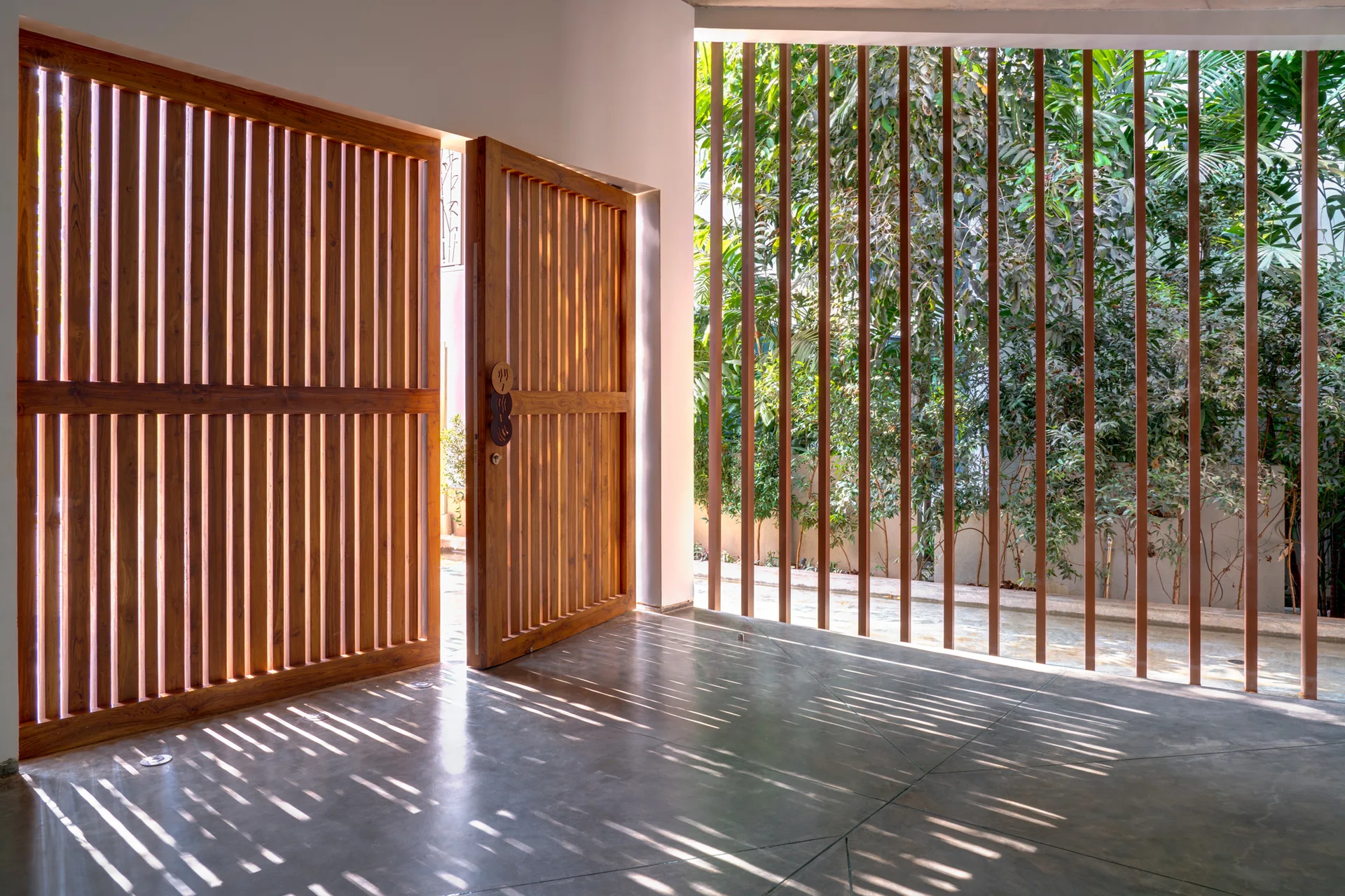 A modern entryway features a large wooden slatted door open to a polished concrete floor with striped sunlight patterns and a green garden view.