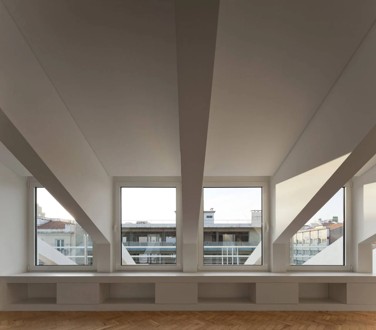 A minimalist attic room features a low white built-in storage bench beneath a row of windows, with a slanted ceiling and herringbone wood floors.