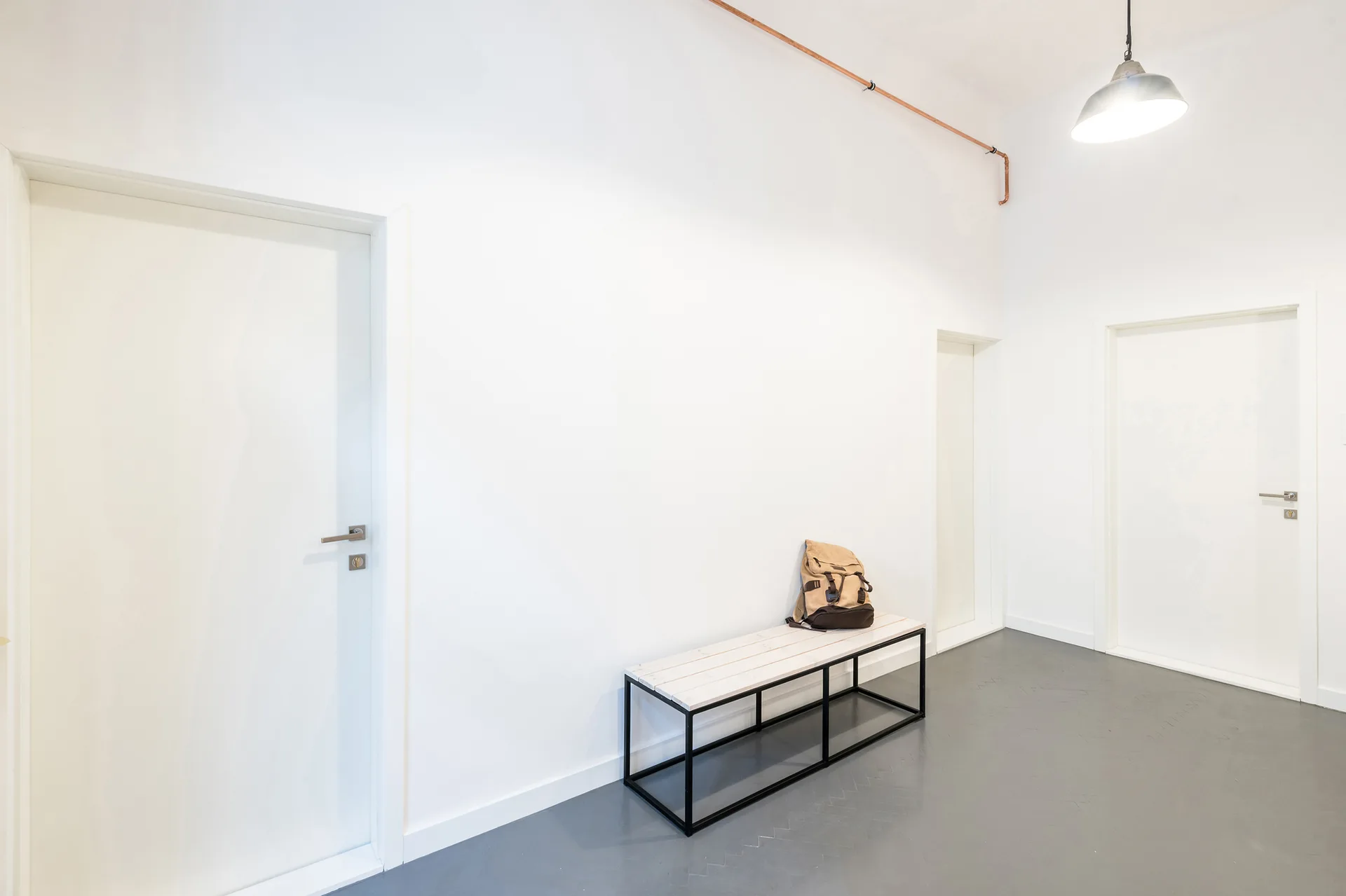 A minimalist white hallway featuring gray floors, multiple doors, a simple wooden bench, and an exposed copper pipe along the wall.