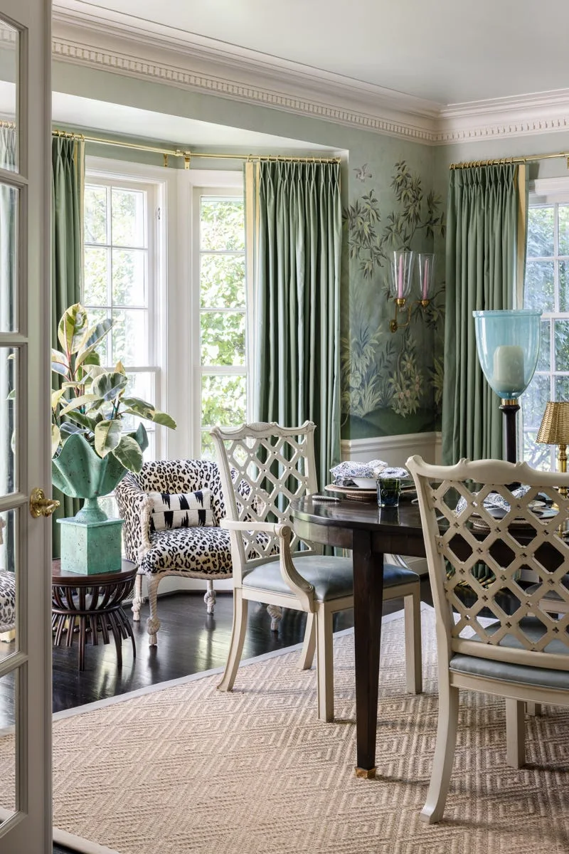 A dining room features a dark wood table and lattice-back chairs on a woven rug, set against scenic green mural wallpaper and matching drapes.