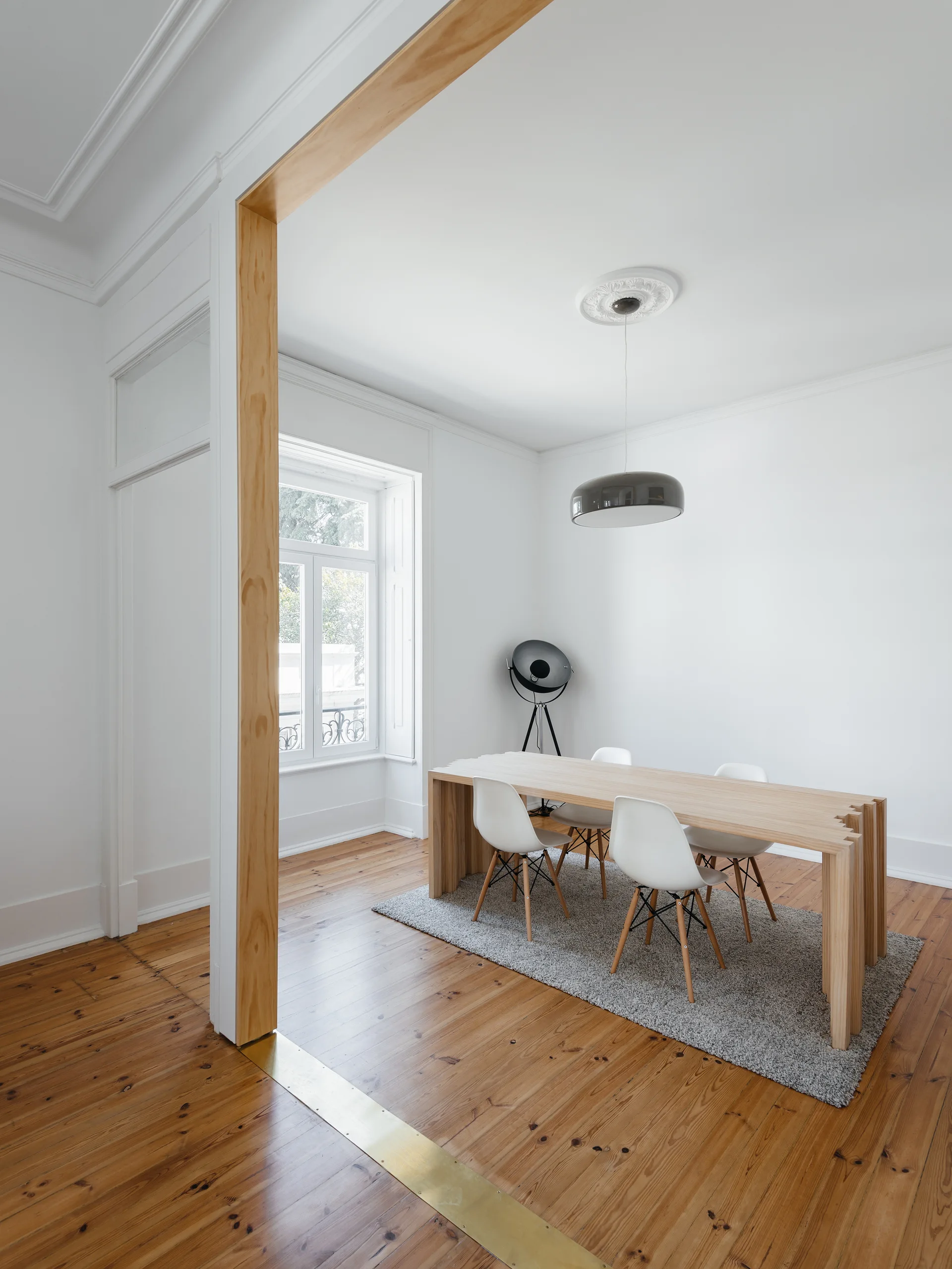 A minimalist dining room features a natural wood table with white chairs on a grey rug, framed by a light wood archway on pine floors.