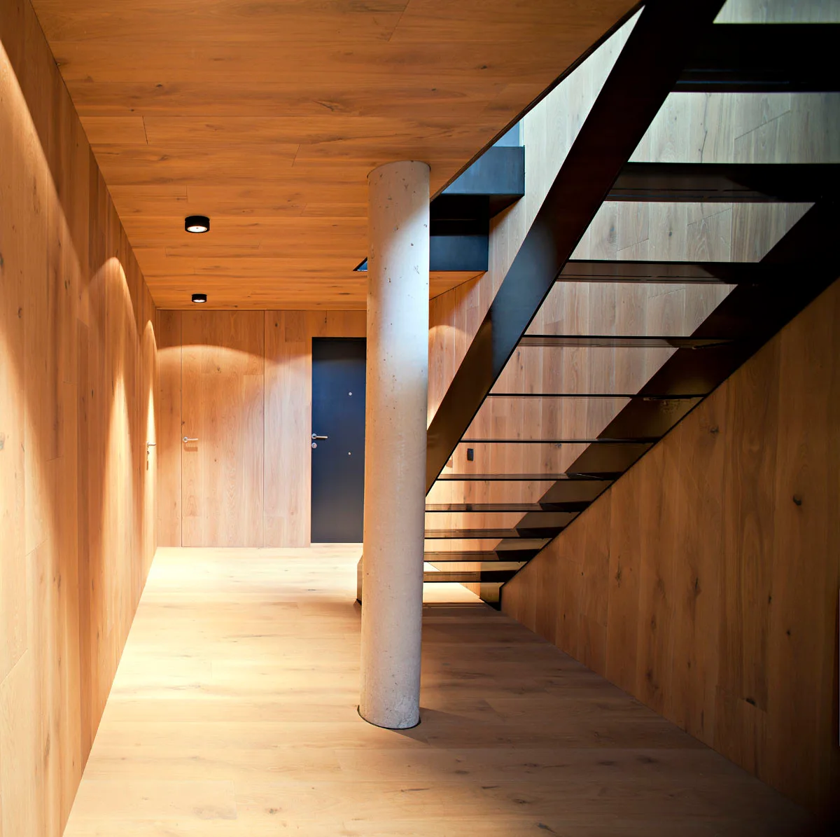 A modern hallway fully clad in warm wood panels features a black open-tread metal staircase and a cylindrical concrete structural column.