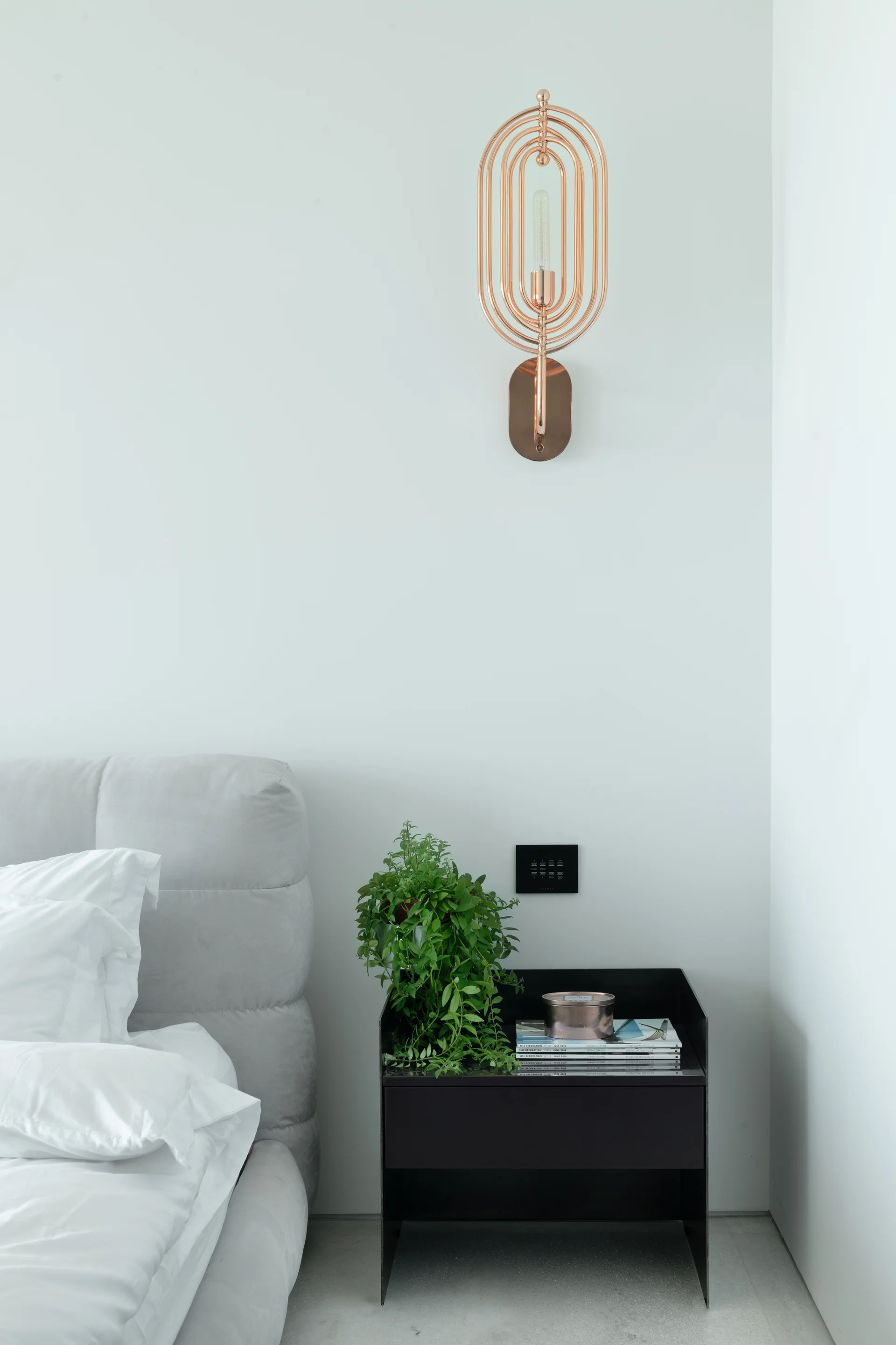 A modern bedroom corner featuring a grey upholstered headboard, a black nightstand with a green plant, and a copper oval wall sconce.