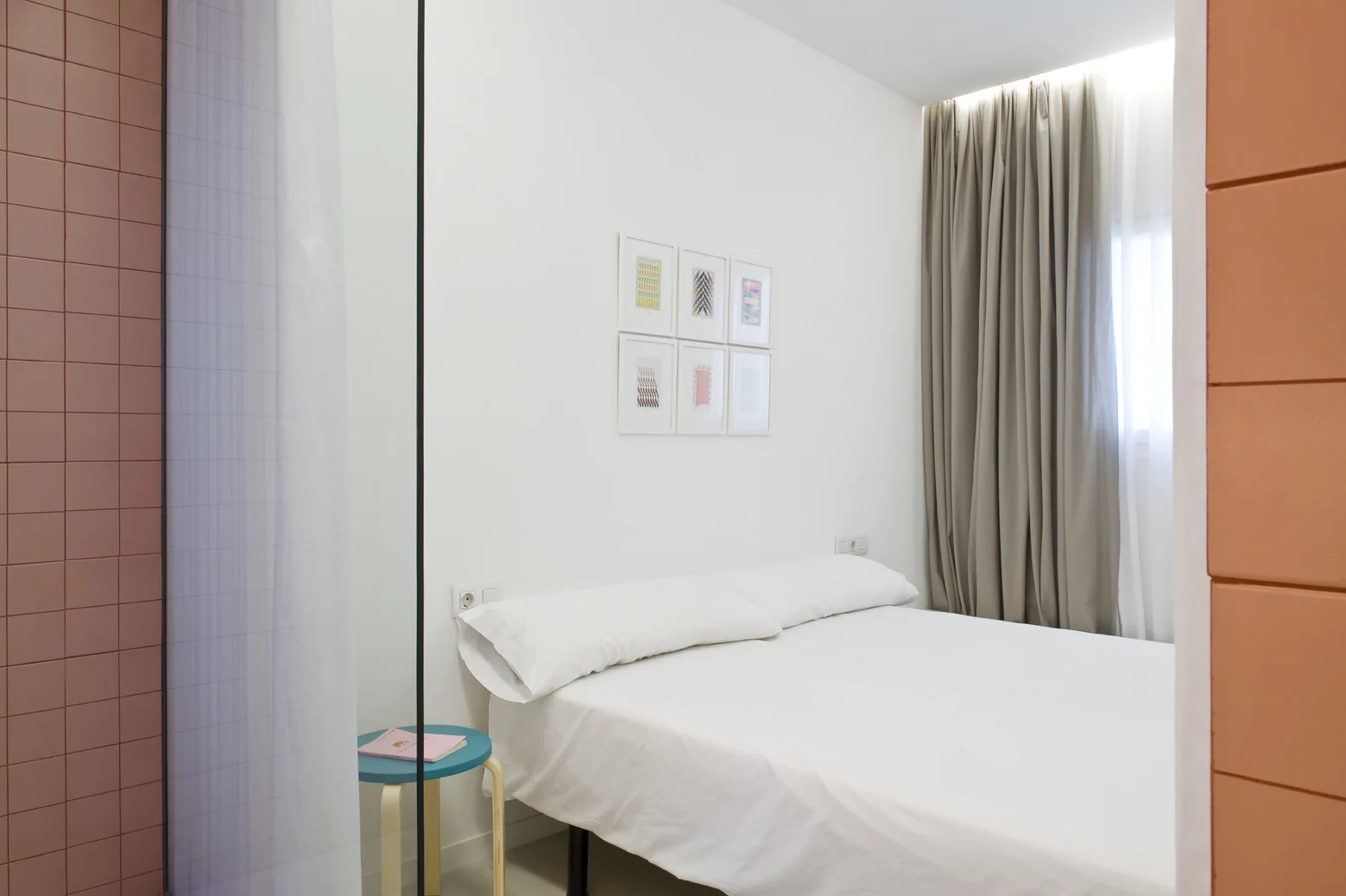 A minimalist bedroom featuring a white bed, grey curtains with cove lighting, a pink tiled partition, and six framed prints on the wall.