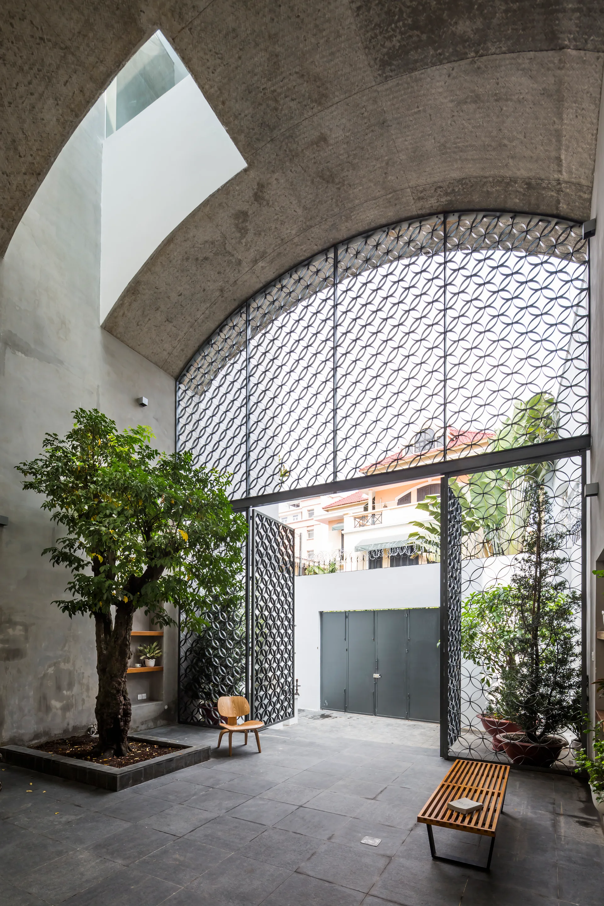 An indoor-outdoor courtyard features a vaulted raw concrete ceiling, a large geometric metal gate, a mature tree, and minimalist wooden furniture.
