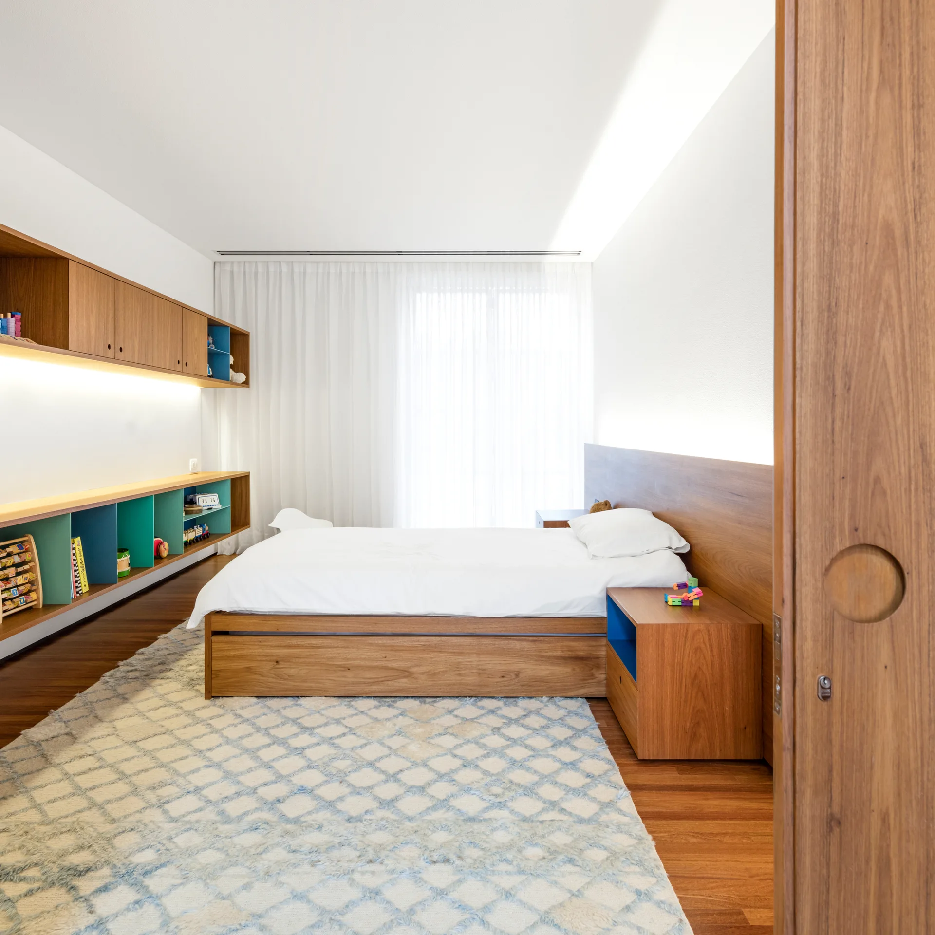 A modern kids bedroom features a wooden platform bed, built-in wall cabinets with teal shelves, and a blue diamond patterned area rug.