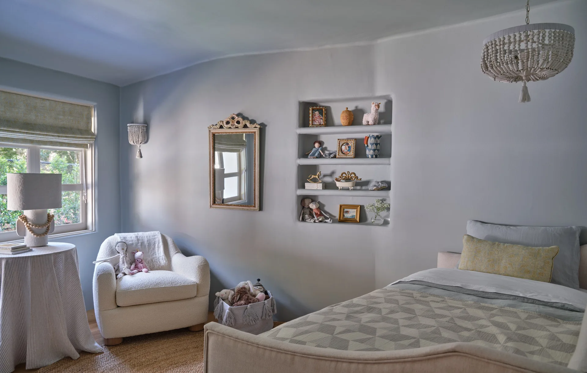 A soft blue children's bedroom features a white boucle armchair, an upholstered bed, an ornate mirror, and built-in plaster wall niches holding toys.