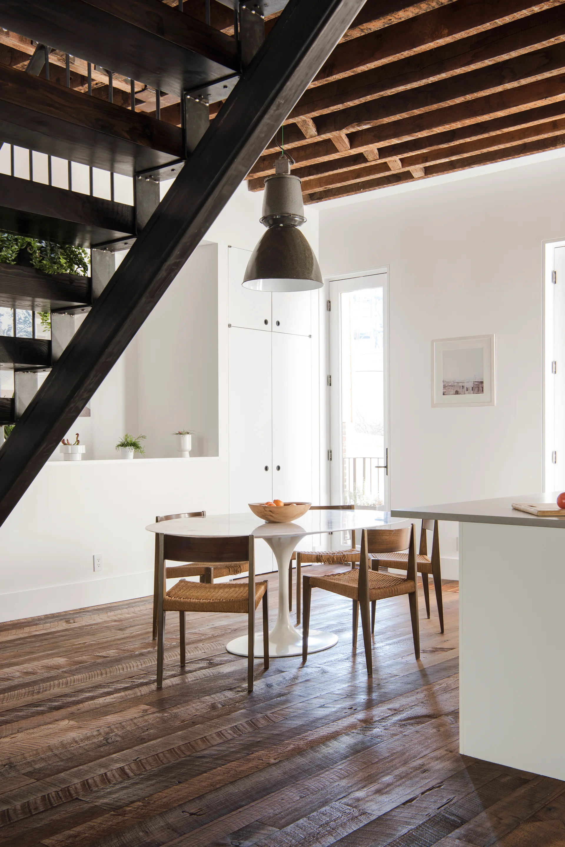 An oval pedestal dining table with wooden rush-seat chairs sits on reclaimed wood floors beneath exposed dark ceiling joists.
