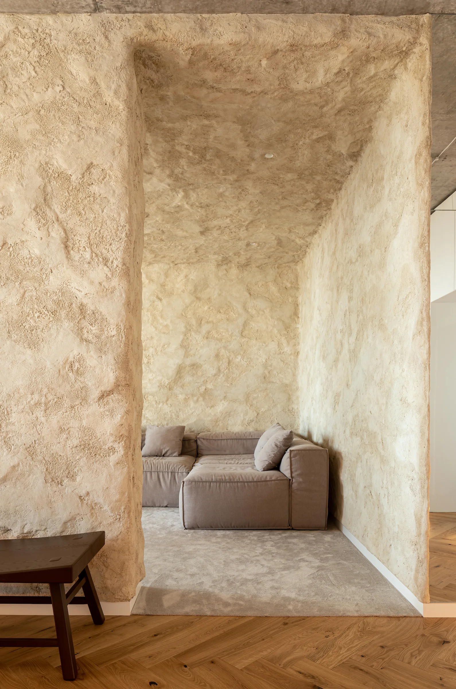 A taupe modular sofa is tucked into a deeply recessed niche with rough beige plaster walls and ceiling, adjacent to herringbone wood floors.