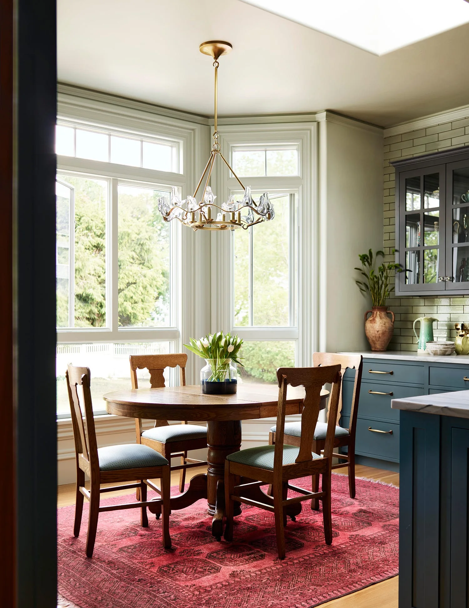 A round wooden table and chairs sit on a red patterned rug in a bright bay window nook next to blue kitchen cabinets.